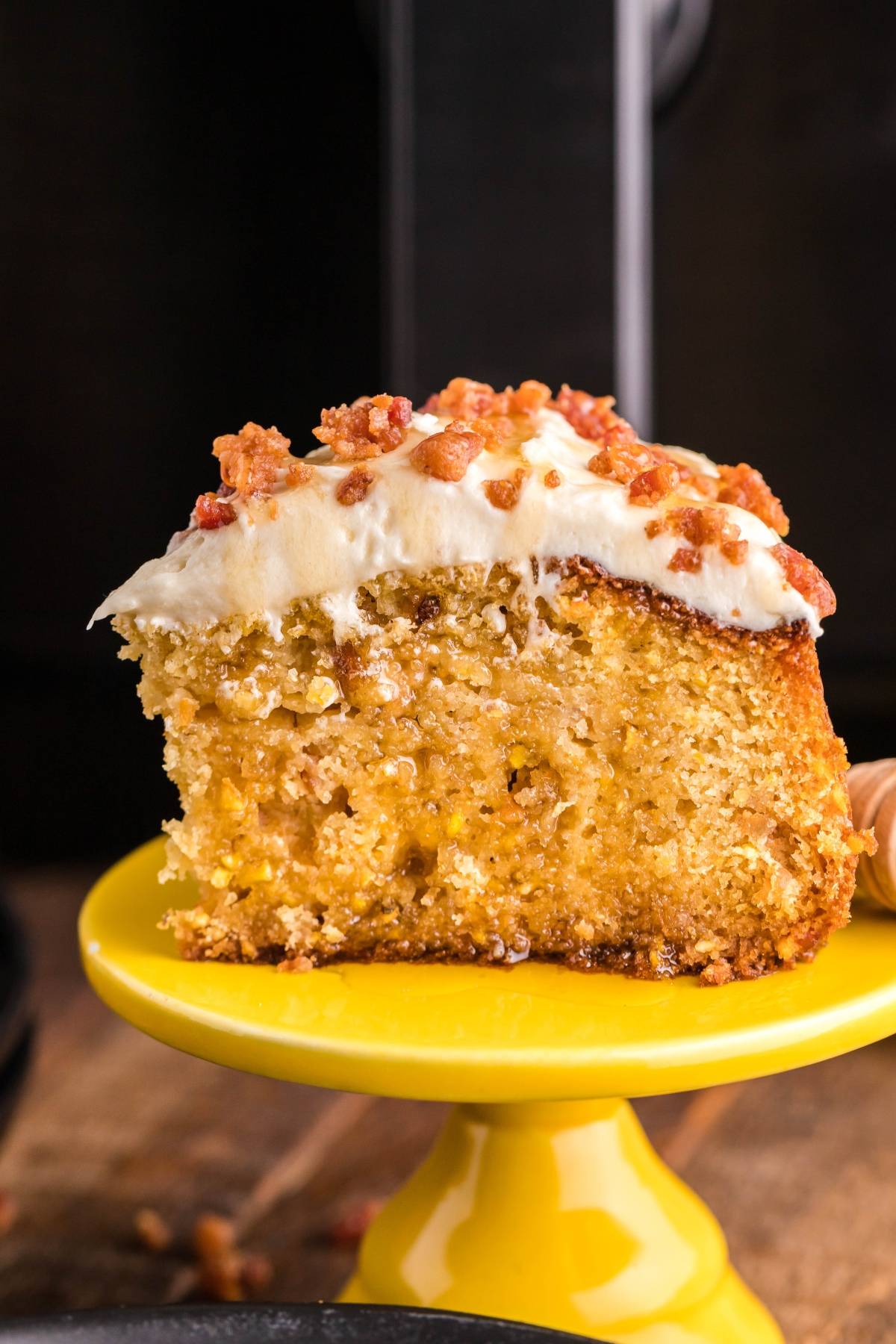 A slice of moist Air Fryer Cornbread Cake with Honey Buttercream frosting and sprinkled nuts sits on a yellow cake stand.