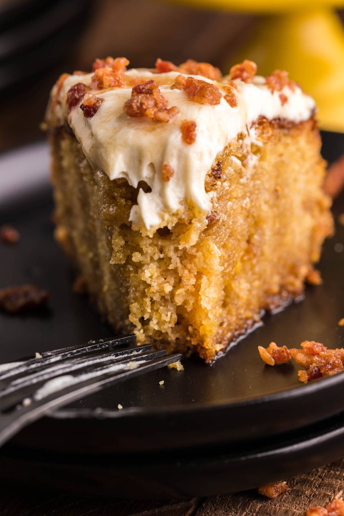 A close-up of moist Air Fryer Cornbread Cake with Honey Buttercream, topped with bacon bits on a black plate, with a fork beside it.