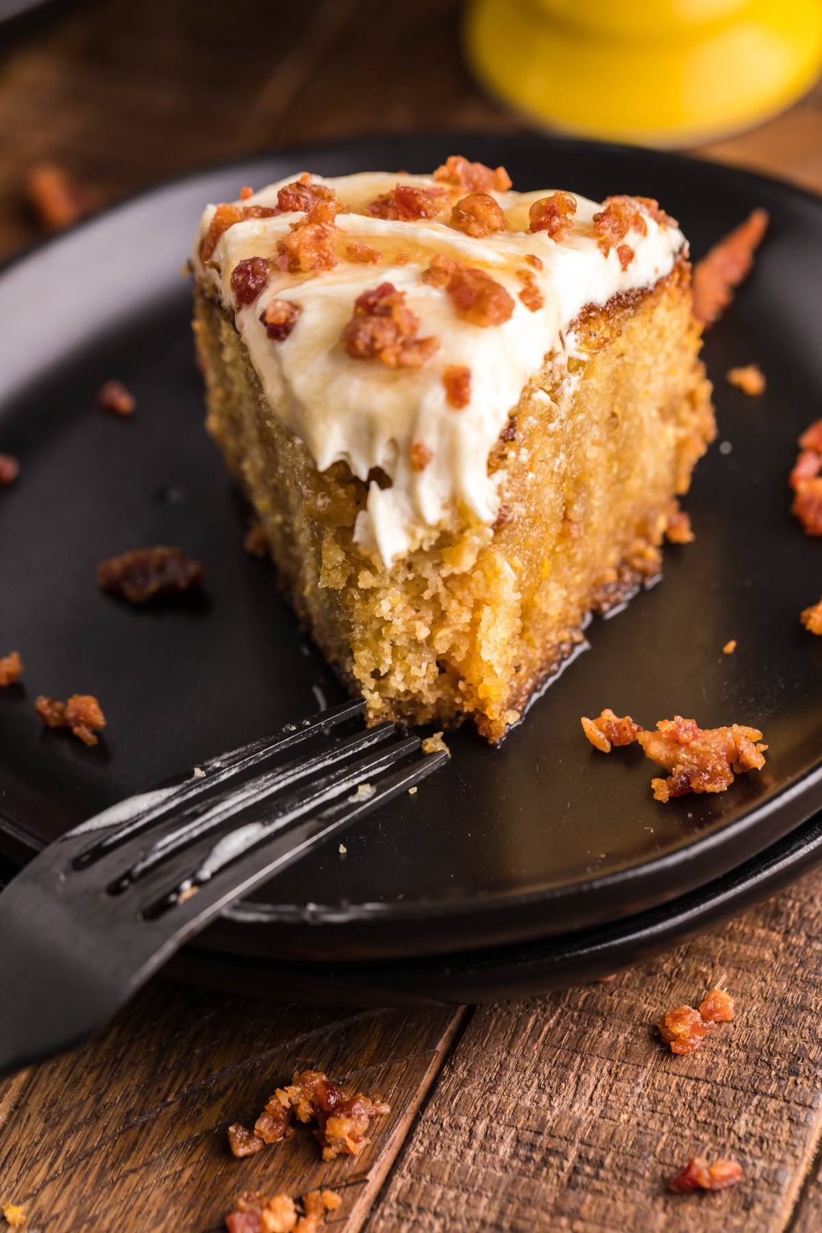 A slice of Air Fryer Cornbread Cake with Honey Buttercream frosting and bacon bits sits on a black plate, with a fork beside it.