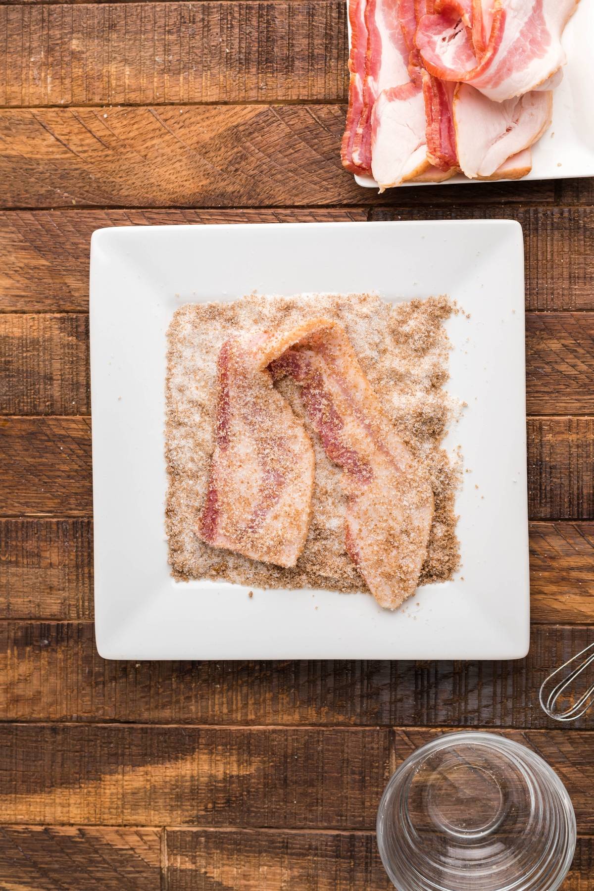 Two raw bacon strips coated in brown sugar on a white square plate, with more bacon on a tray nearby—perfect alongside sweet cornbread with icing for an irresistible sweet and savory pairing.