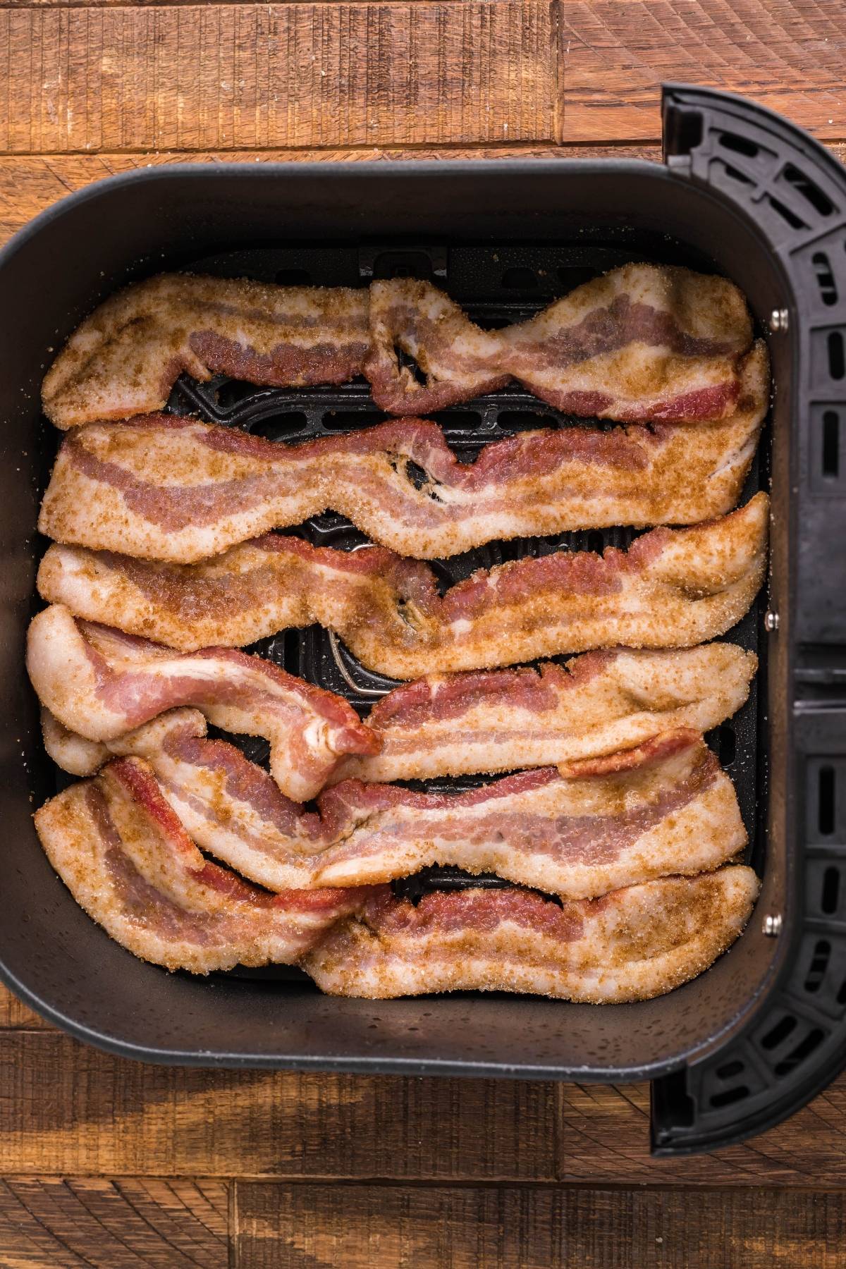 Raw bacon strips seasoned with spices, arranged in a single layer inside an air fryer basket—perfect alongside a slice of Air Fryer Cornbread Cake with Honey Buttercream.