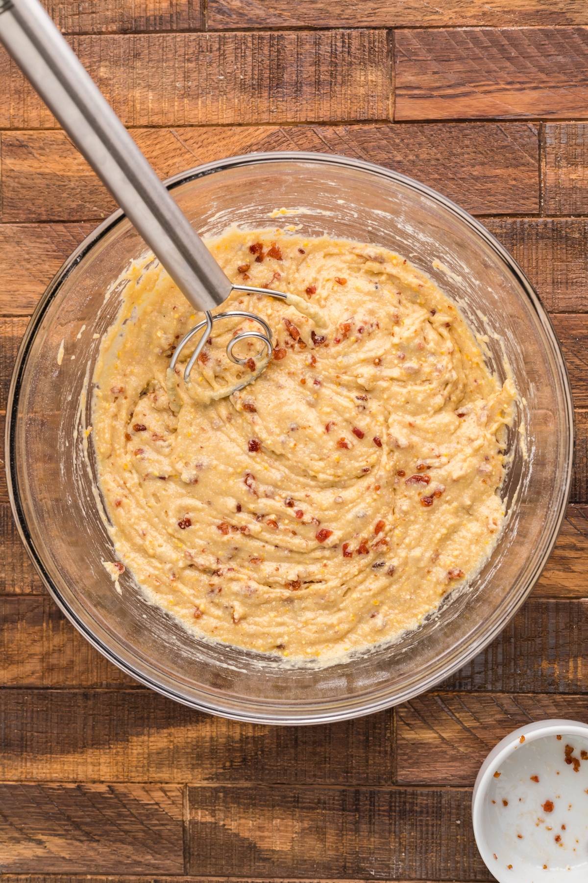 A glass bowl of muffin batter with bits mixed in and a whisk resting inside, on a wooden surface—perfect for an air fryer cornbread recipe or sweet cornbread with icing.