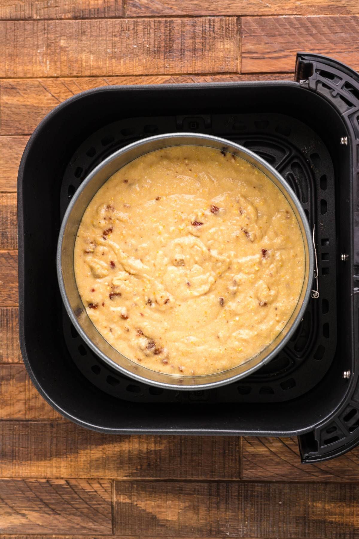 Cake batter for Air Fryer Cornbread Cake with Honey Buttercream frosting sits in a round pan inside an air fryer basket on a wooden countertop.
