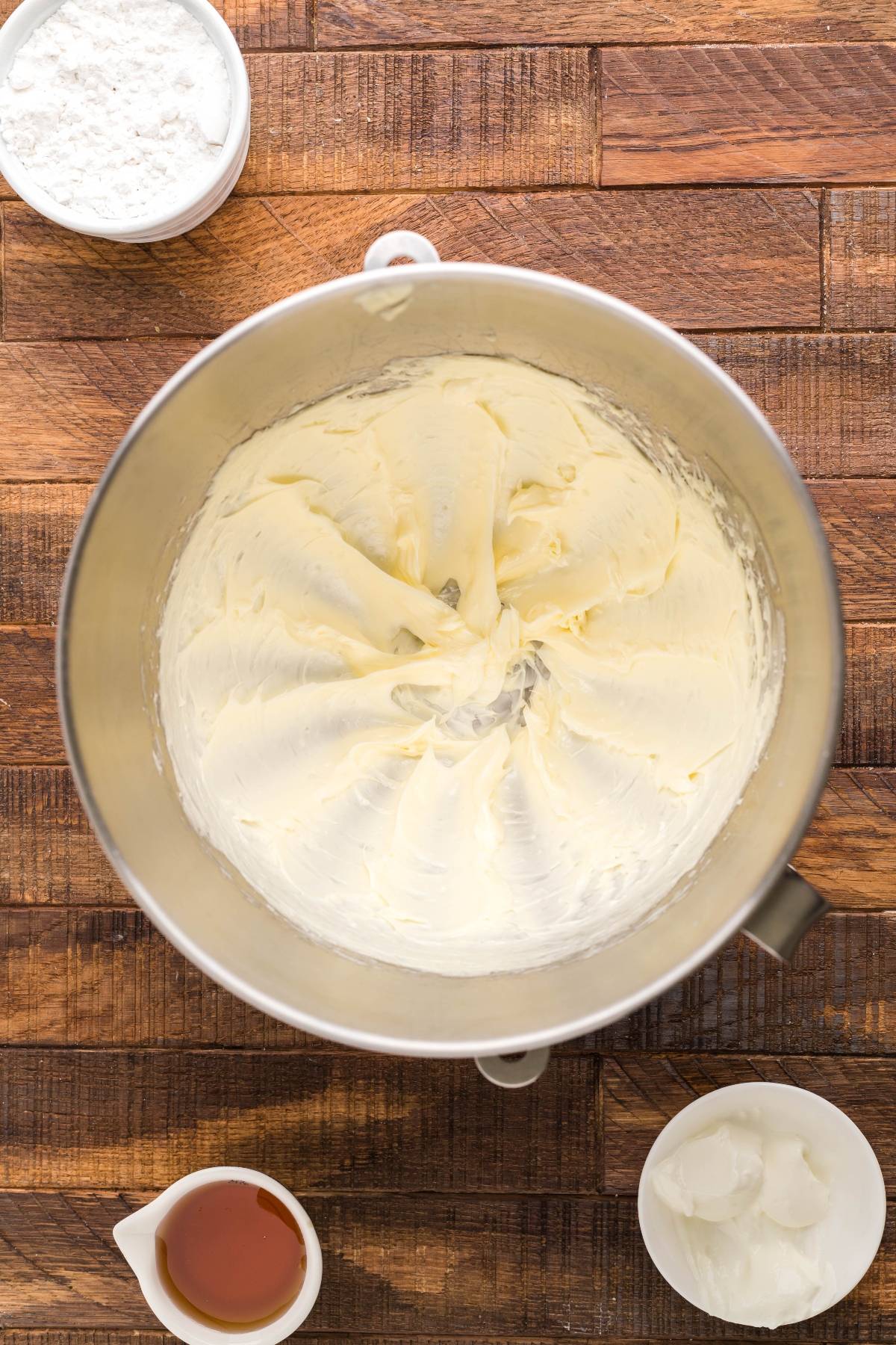 A mixing bowl with creamy Honey Buttercream frosting, surrounded by bowls of powdered sugar, shortening, and vanilla extract—perfect for topping your Air Fryer Cornbread Cake with Honey Buttercream.