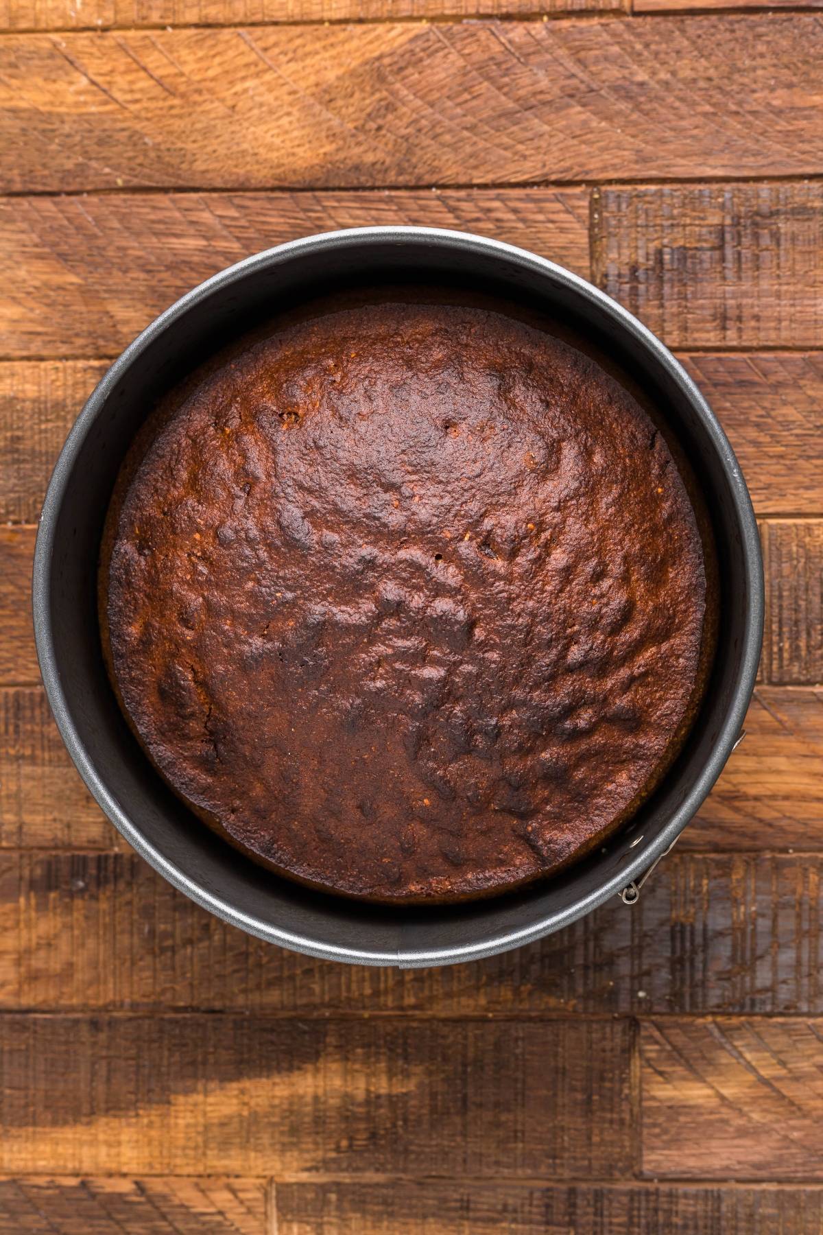 A round, baked chocolate cake in a metal pan on a wooden surface, topped with luscious Honey Buttercream frosting.