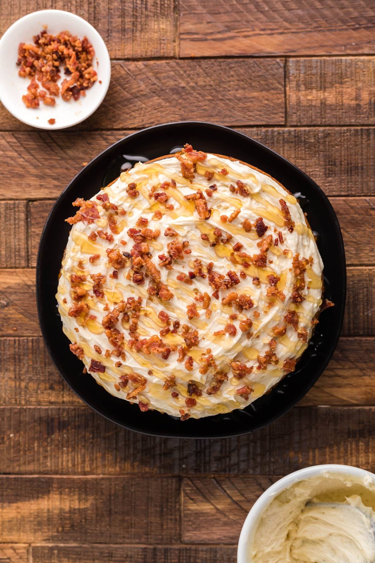 Round cornbread dessert with white Honey Buttercream frosting, caramel drizzle, and bacon bits on a black plate, on a wooden table.