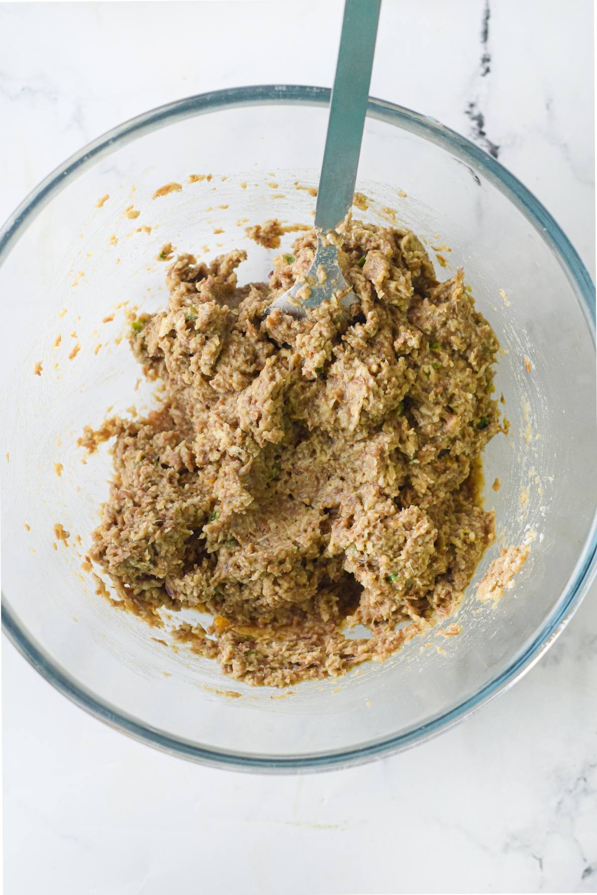 A glass bowl with a mixture of raw vegan meatball recipe ingredients and a fork sits on a marble surface, ready to be transformed into delicious vegan meatballs with spaghetti marinara.