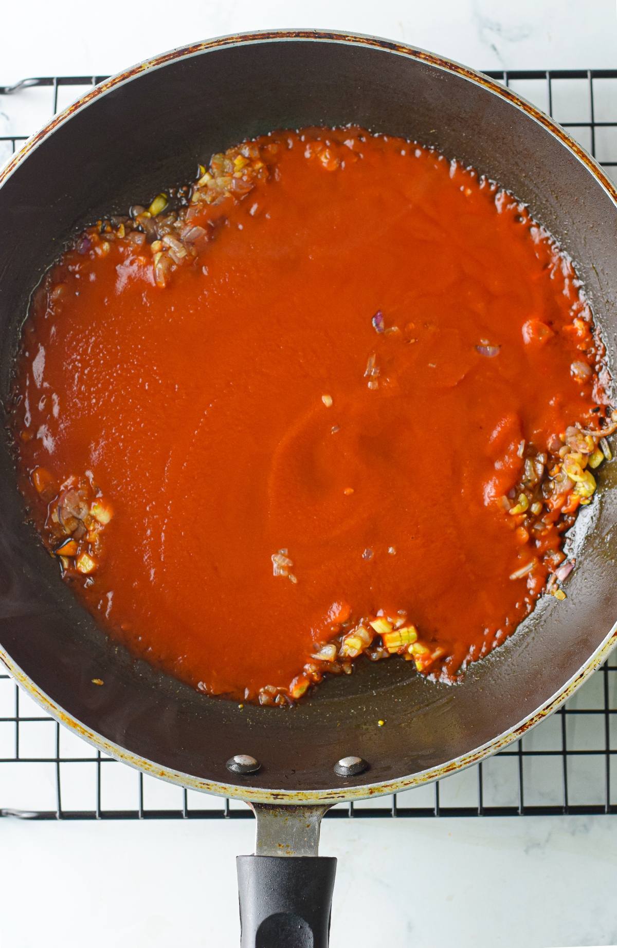 A frying pan with tomato sauce and sautéed onions, perfect for preparing vegan meatballs with spaghetti marinara, rests on a cooling rack.