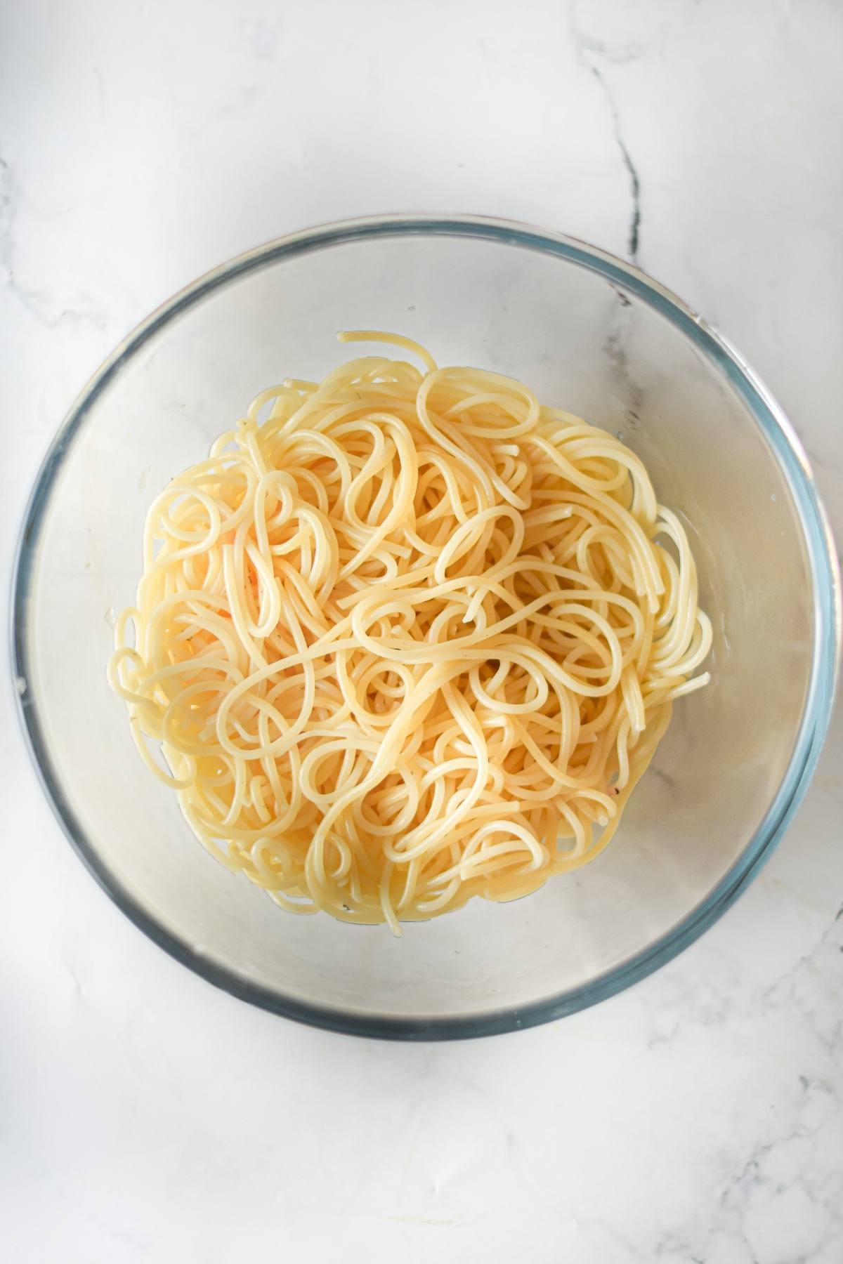 A clear glass bowl filled with plain cooked spaghetti noodles on a white marble surface, ready to be paired with your favorite easy vegan meatballs.