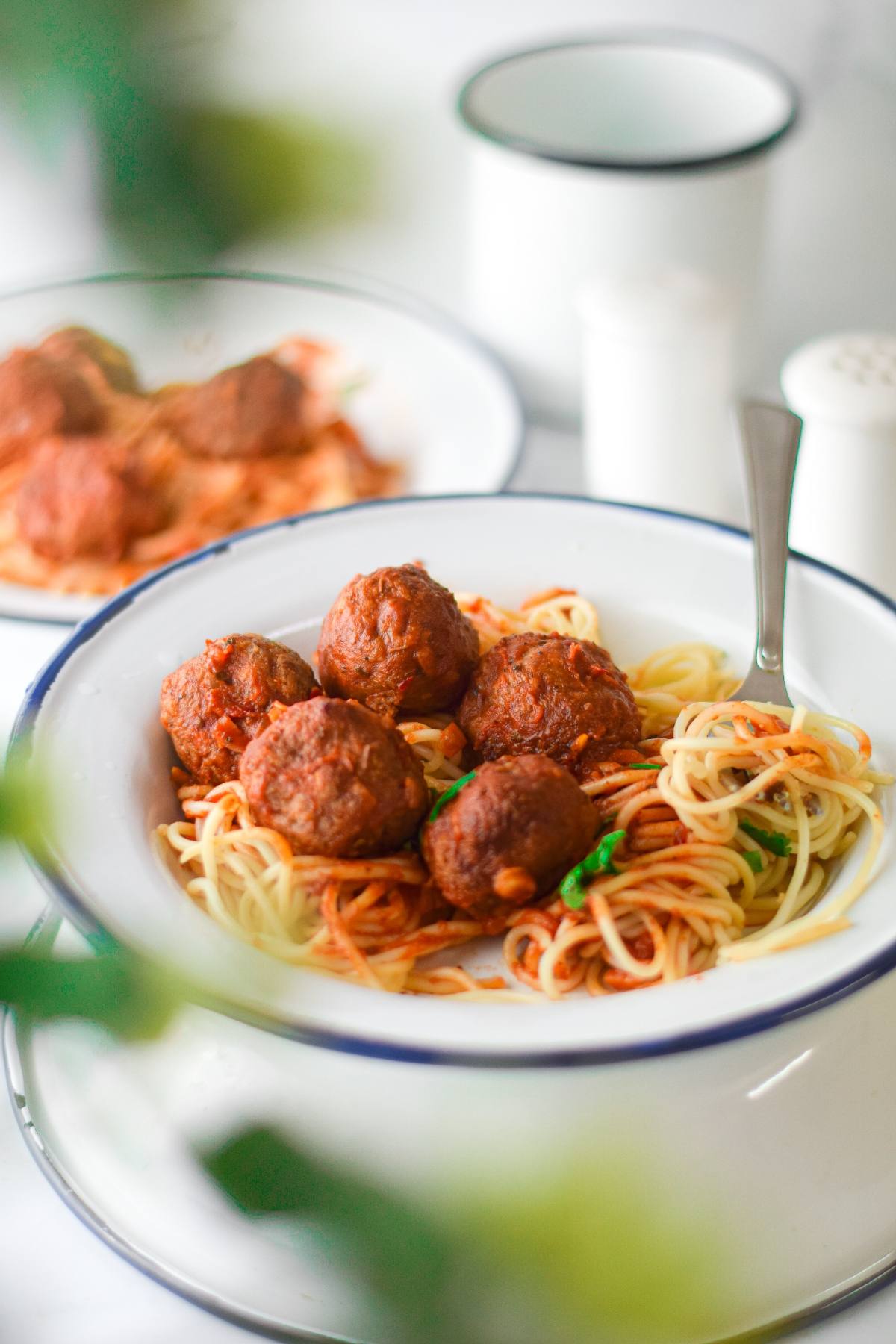 A bowl of spaghetti with rich tomato sauce and vegan meatballs with spaghetti marinara, accompanied by a fork and a blurred plate in the background.