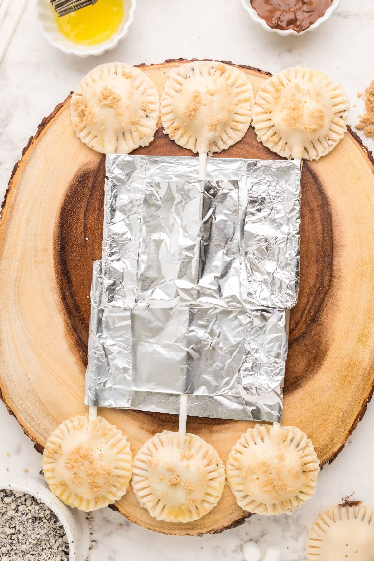 Six unbaked air fryer s’more pie pops on sticks are arranged around foil on a wooden board, ready for baking—a fun summer BBQ dessert idea.