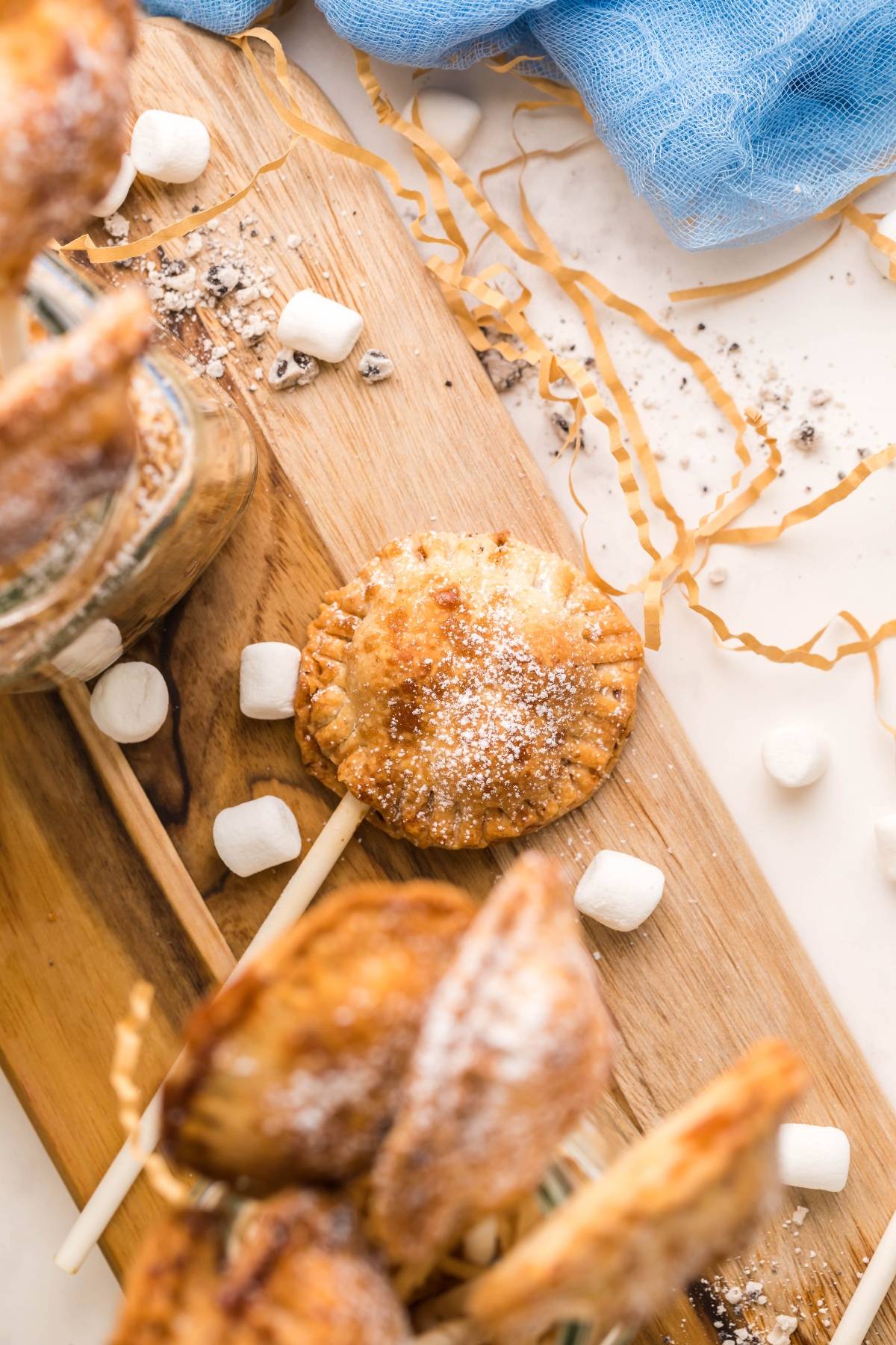 A round, golden-brown pastry on a stick, dusted with powdered sugar and surrounded by mini marshmallows—these air fryer s’more pie pops are the ultimate summer BBQ dessert idea.