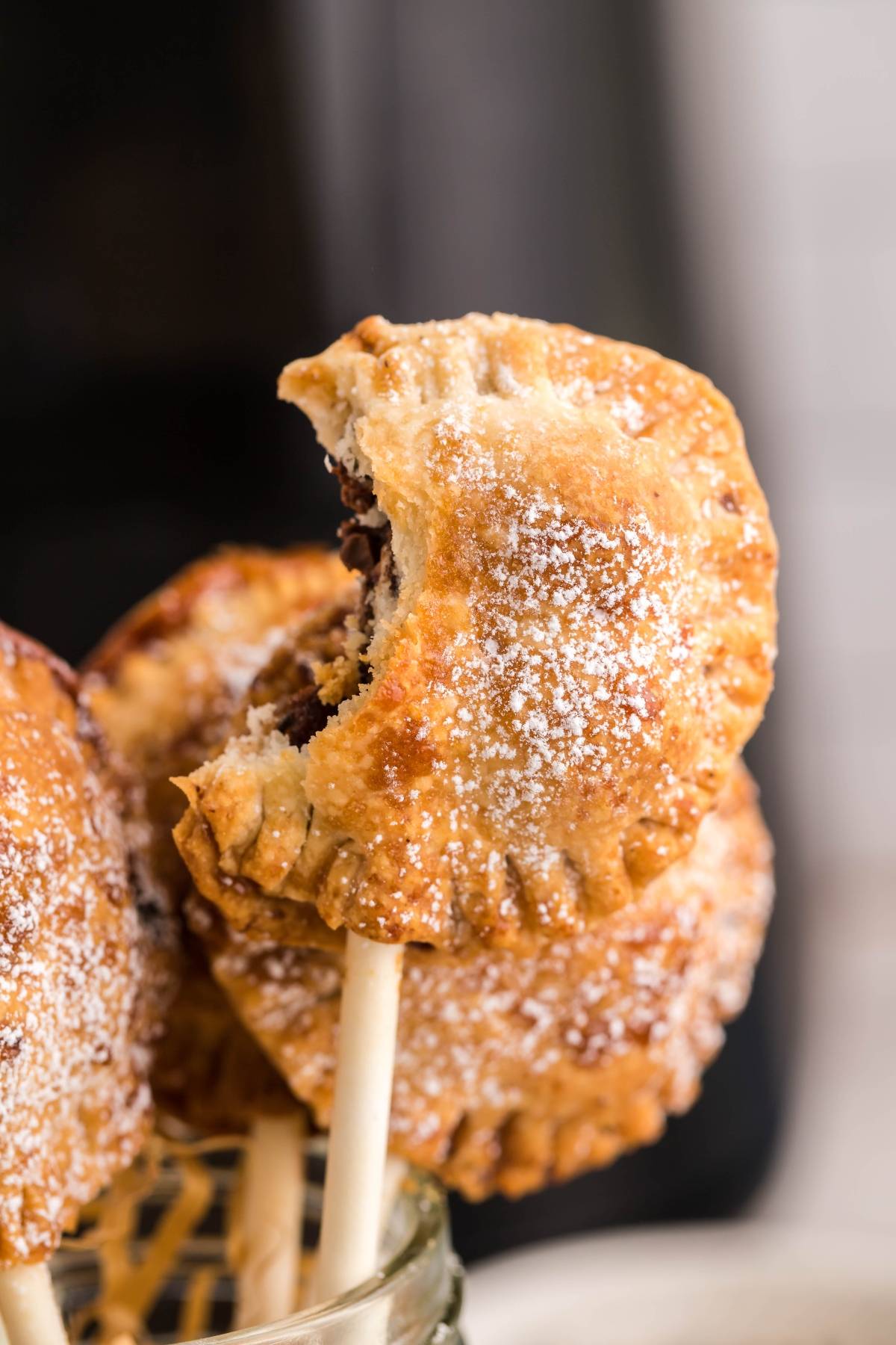 A closeup of a s’mores mini pie on a stick with a bite taken out, dusted with powdered sugar—an easy s’more treat and perfect summer BBQ dessert idea.