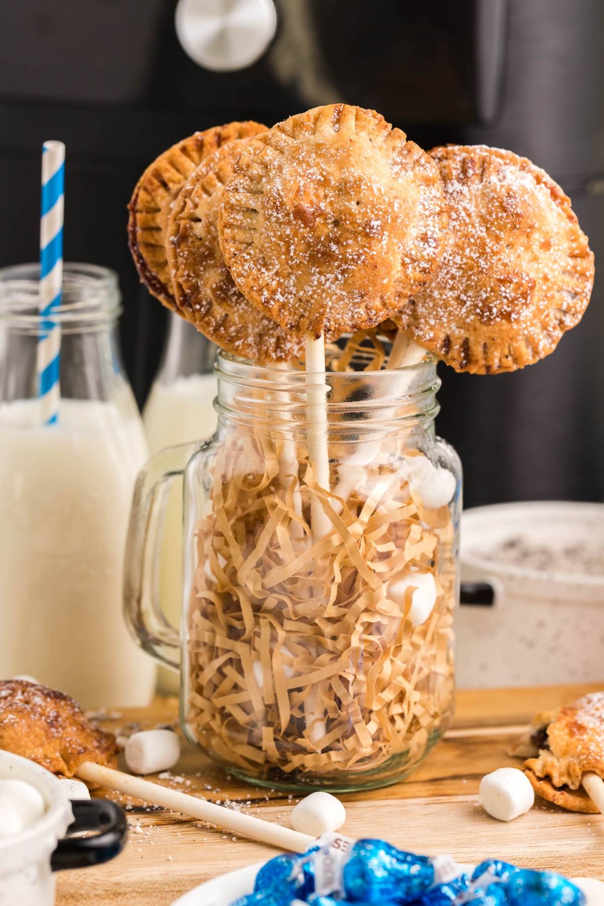 Round pie pops on sticks are displayed in a jar with shredded paper, surrounded by bottles of milk—a fun and easy s’more treats display perfect for your summer BBQ dessert idea.