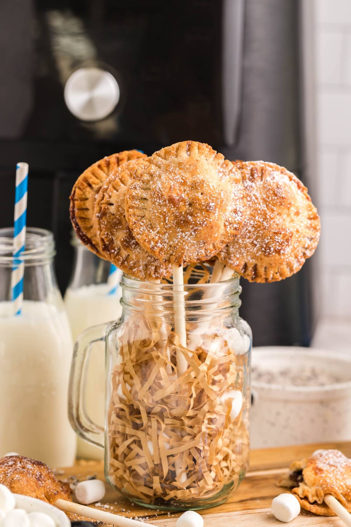 Round pie pops on sticks in a mason jar, with bottles of milk and straws in the background—these easy s’more treats are perfect for sharing.