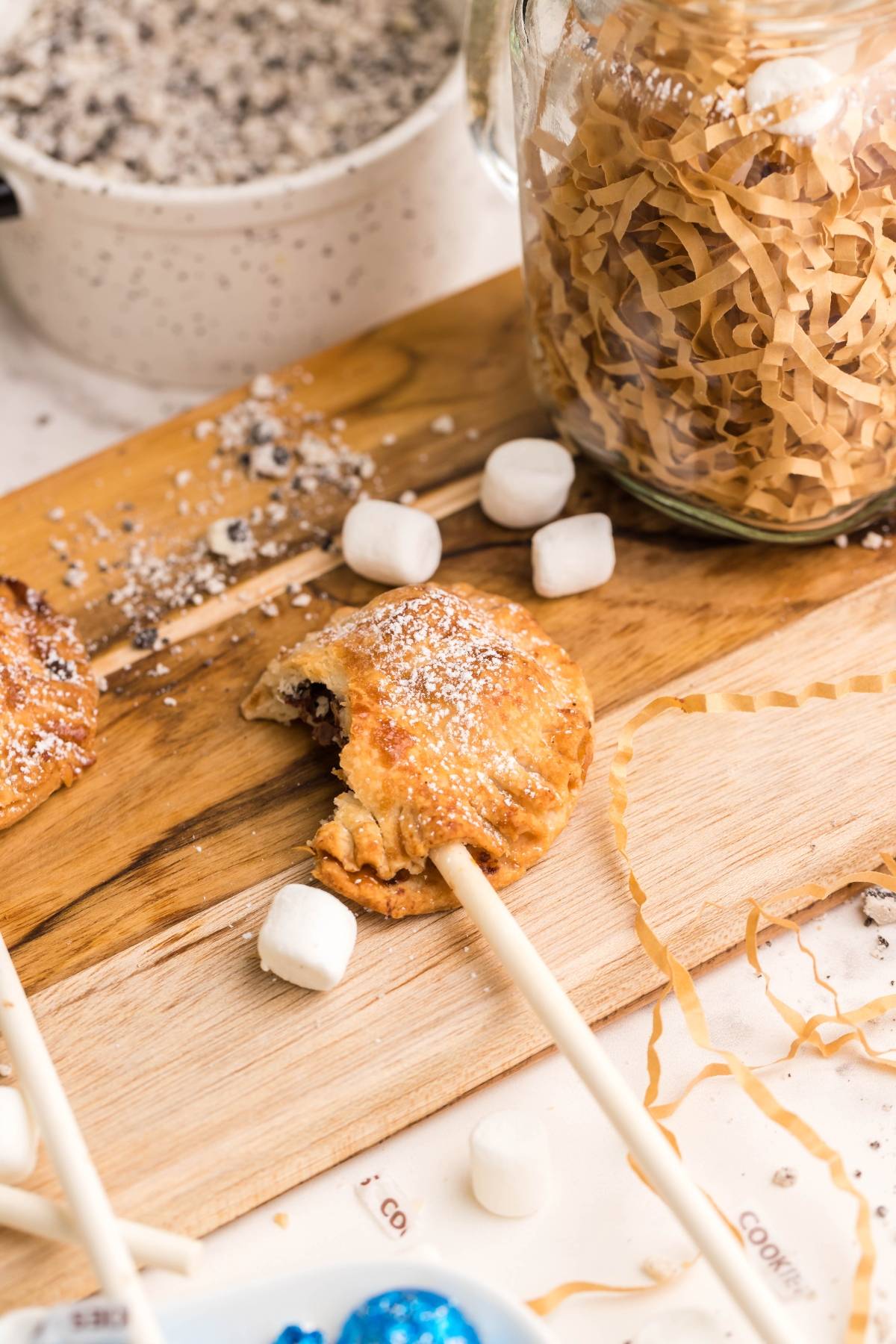 A chocolate-filled pastry on a stick with a bite taken out, surrounded by marshmallows and shredded paper—these air fryer s’more pie pops are the perfect easy s’more treats for any occasion.