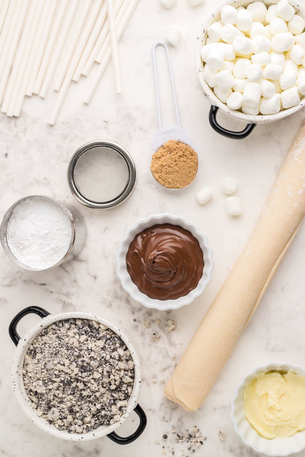 Baking ingredients on a marble surface: perfect for an air fryer s’more recipe. Includes marshmallows, chocolate, cookie crumbs, dough, butter, and lollipop sticks—a fun summer BBQ dessert idea!.