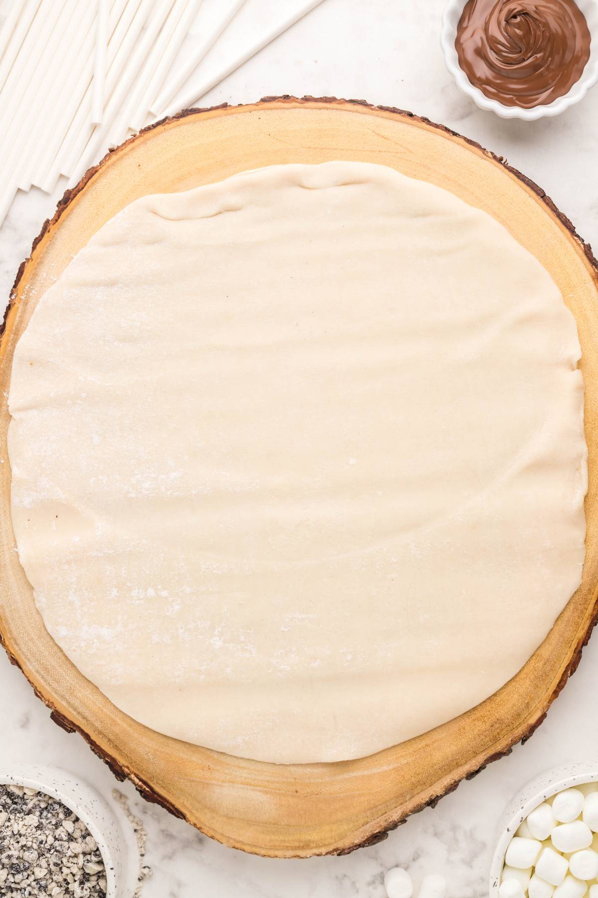 Round sheet of dough on a wooden board, surrounded by Nutella, marshmallows, sprinkles, and sticks—perfect for creating s’mores mini pies as a fun summer BBQ dessert idea.