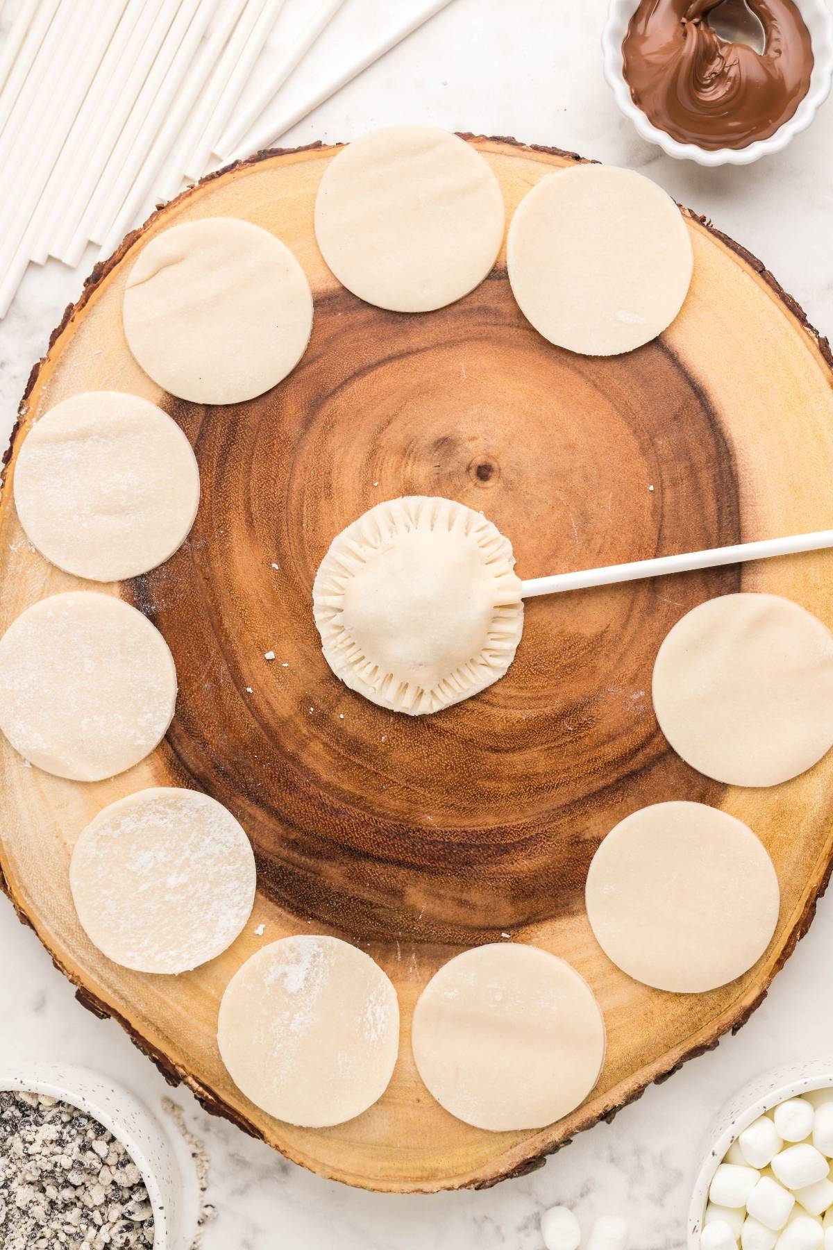 Round pastry dough pieces arranged in a circle on a wood board, with one filled and sealed on a stick—perfect for easy s’more treats like air fryer s’more pie pops.