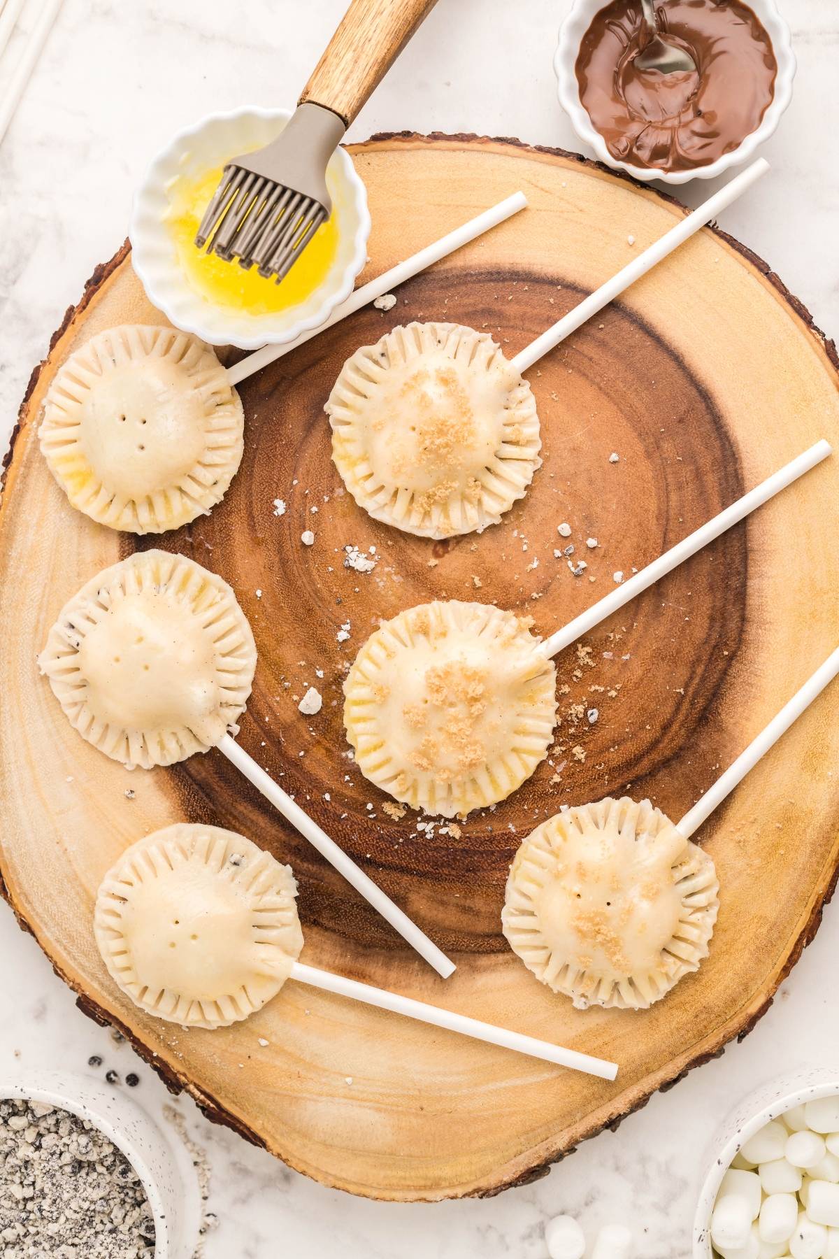 Six unbaked air fryer s’more pie pops on sticks rest on a wooden board, with bowls of egg wash and melted chocolate nearby—a fun, easy s’more treat and perfect summer BBQ dessert idea.