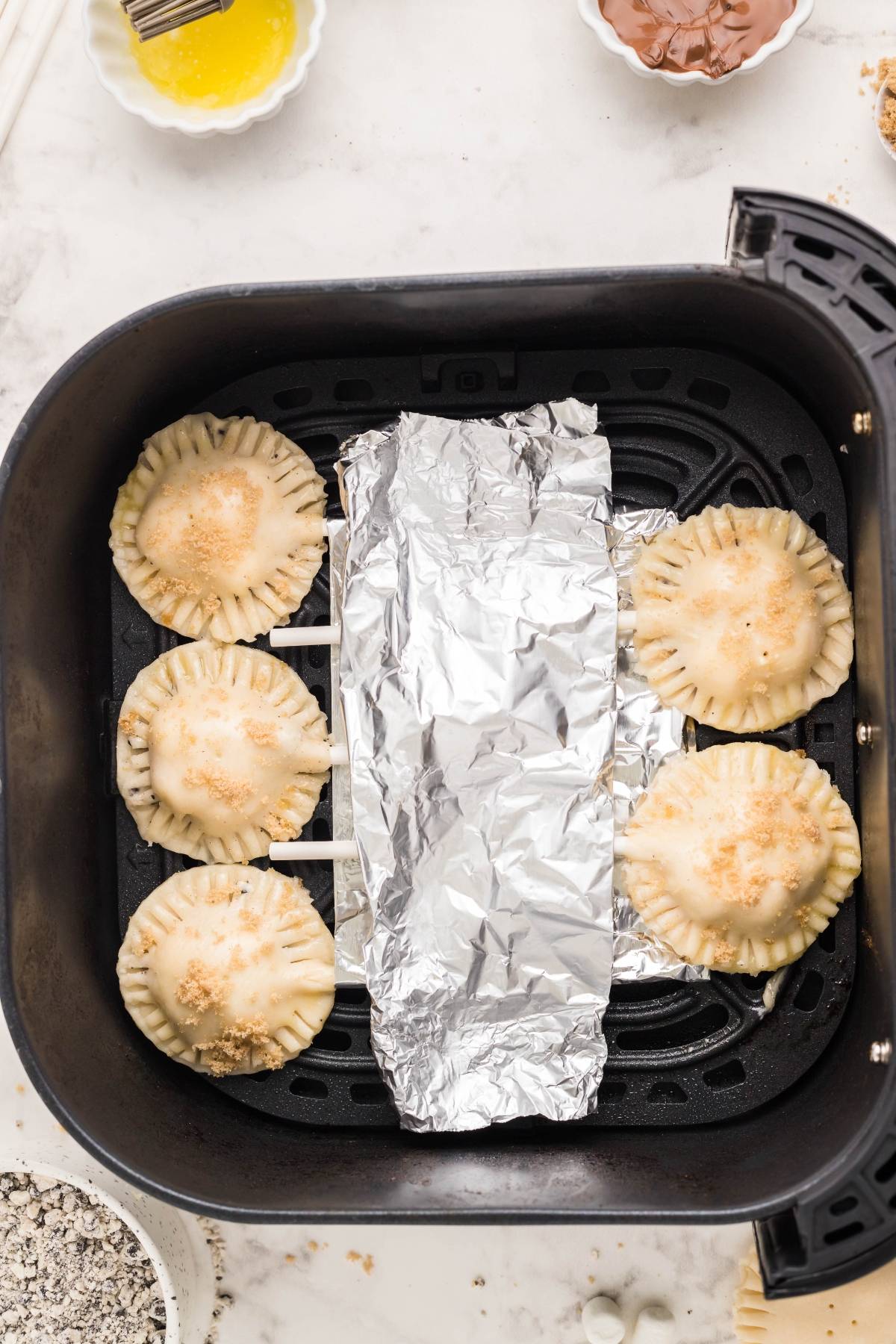 Five round s’mores mini pies with crimped edges sit in an air fryer basket, one side covered with a piece of foil—an easy s’more treat perfect for dessert lovers.