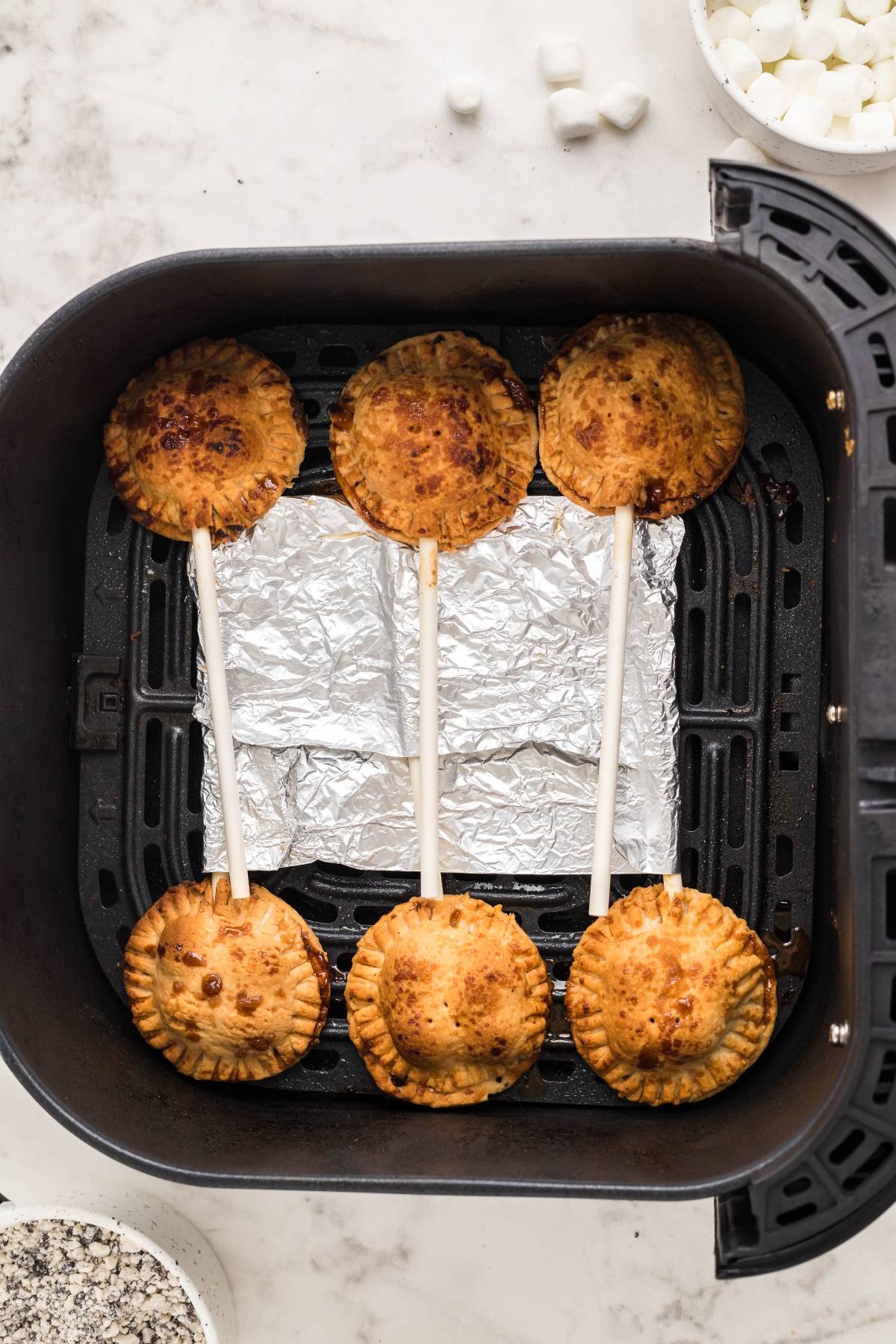 Five air fryer s’more pie pops on sticks in a foil-lined air fryer basket, surrounded by marshmallows—an easy s’more treat for any occasion.