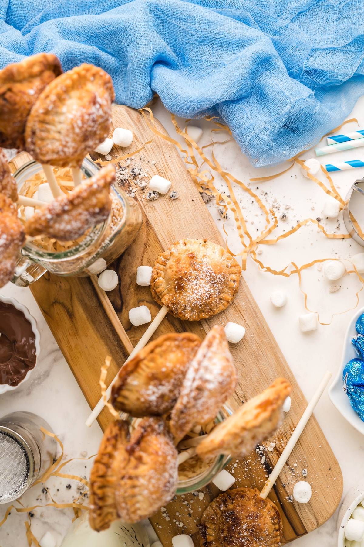 Mini pie pops on sticks, dusted with powdered sugar and surrounded by mini marshmallows on a wooden board—a fun summer BBQ dessert idea inspired by an air fryer s’more recipe.