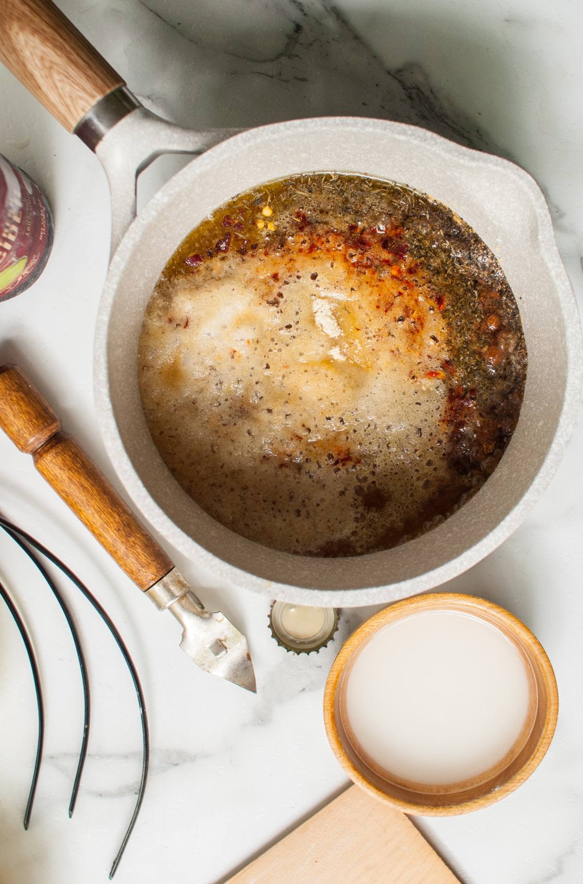 A saucepan with simmering brown liquid, wooden utensils, a bottle cap, and a small bowl of white liquid nearby—perfect for preparing an apple cider chicken marinade for delicious Apple Cider Chicken Skewers with Gouda Sauce.