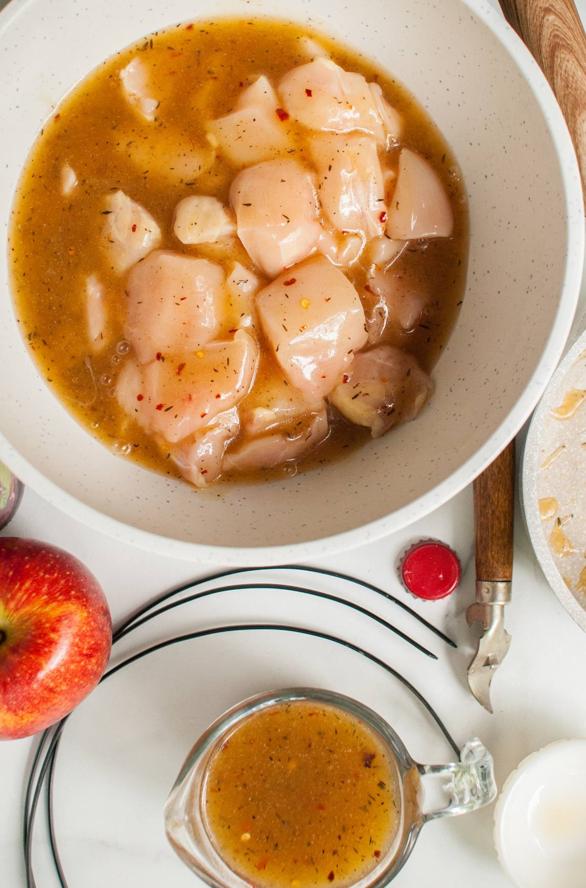 Diced raw chicken marinating in a seasoned sauce, perfect for creating cider grilled chicken skewers, with an apple and extra marinade nearby on a white surface.