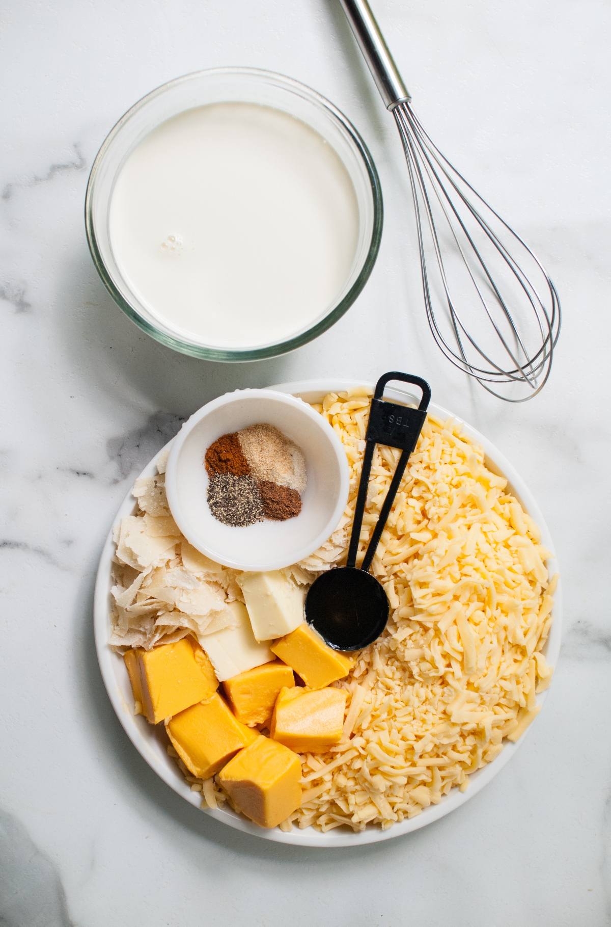 A plate with shredded cheese, butter, spices, and a bowl of milk next to a whisk on a marble surface—perfect for crafting Gouda sauce to pair with Apple Cider Chicken Skewers or your favorite apple cider chicken marinade.