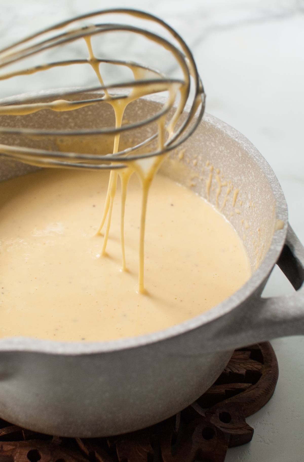 Whisk lifting creamy Gouda Cheese Sauce from a saucepan on a white countertop, perfect for drizzling over Apple Cider Chicken Skewers with Gouda Sauce.