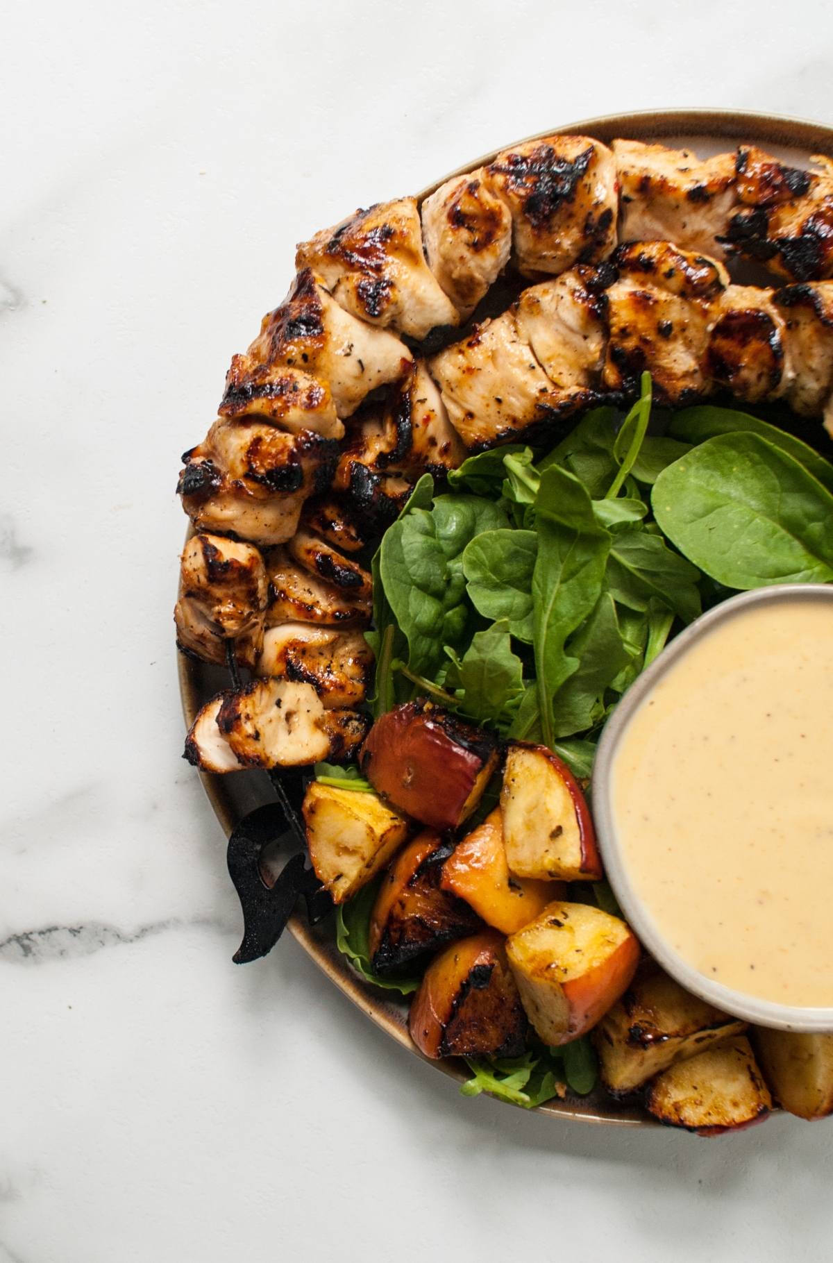 A plate with Apple Cider Chicken Skewers with Gouda Sauce, fresh spinach, roasted vegetables, and a bowl of creamy dipping sauce.