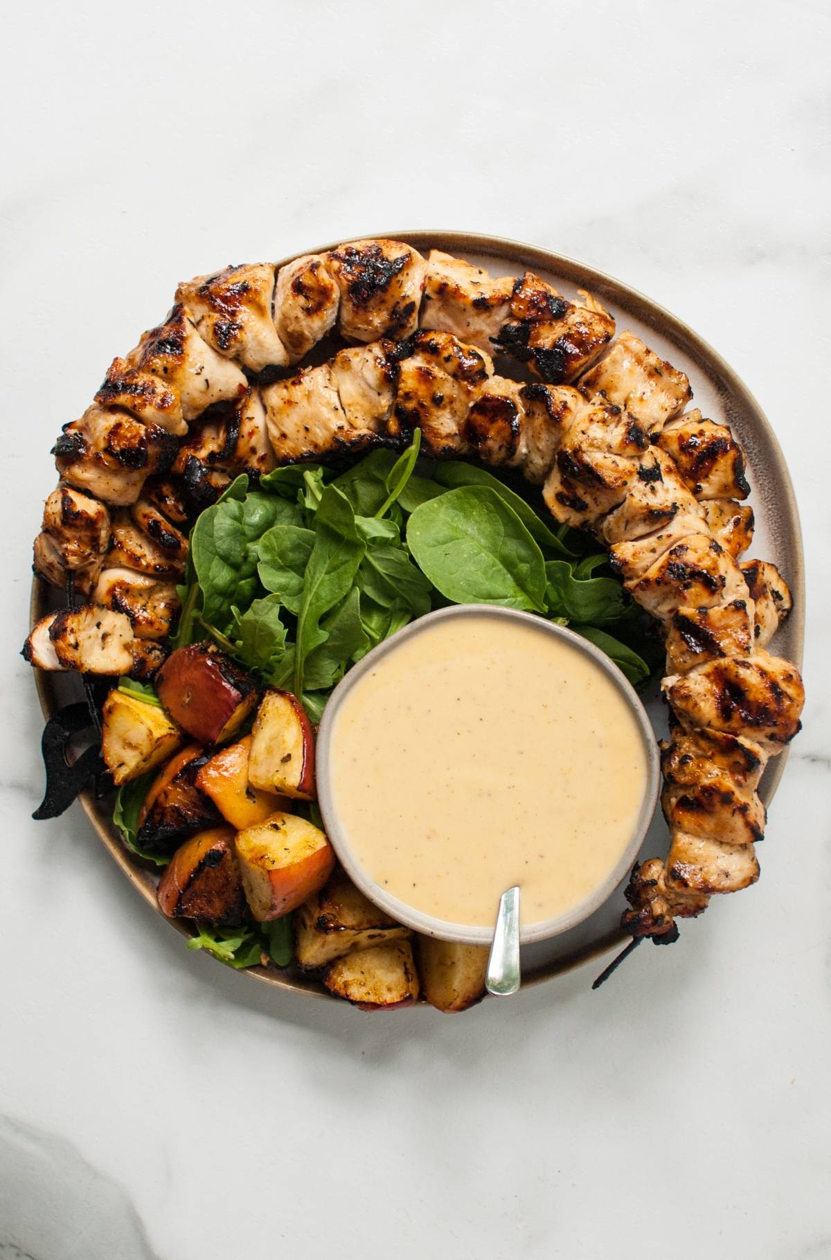 A plate with cider grilled chicken skewers, roasted potatoes, fresh greens, and a bowl of creamy dipping sauce.