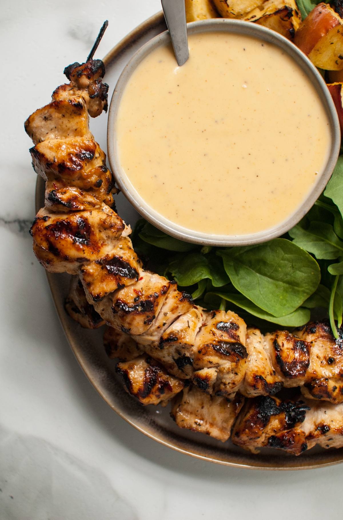 Grilled cider chicken skewer on a plate with spinach and a small bowl of creamy Gouda sauce for dipping.