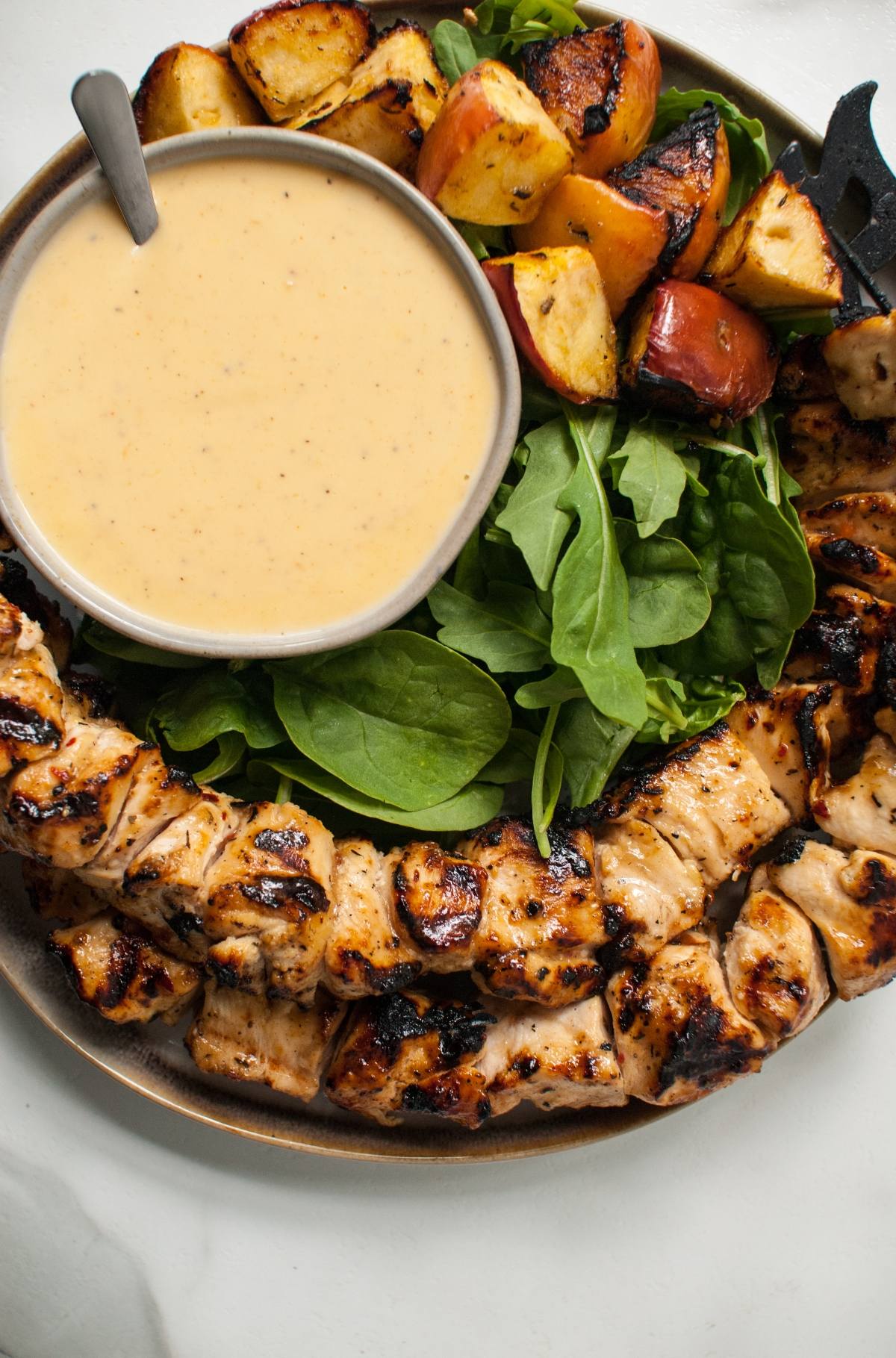 A plate with apple cider chicken skewers with Gouda sauce, roasted potatoes, fresh greens, and a bowl of dipping sauce.