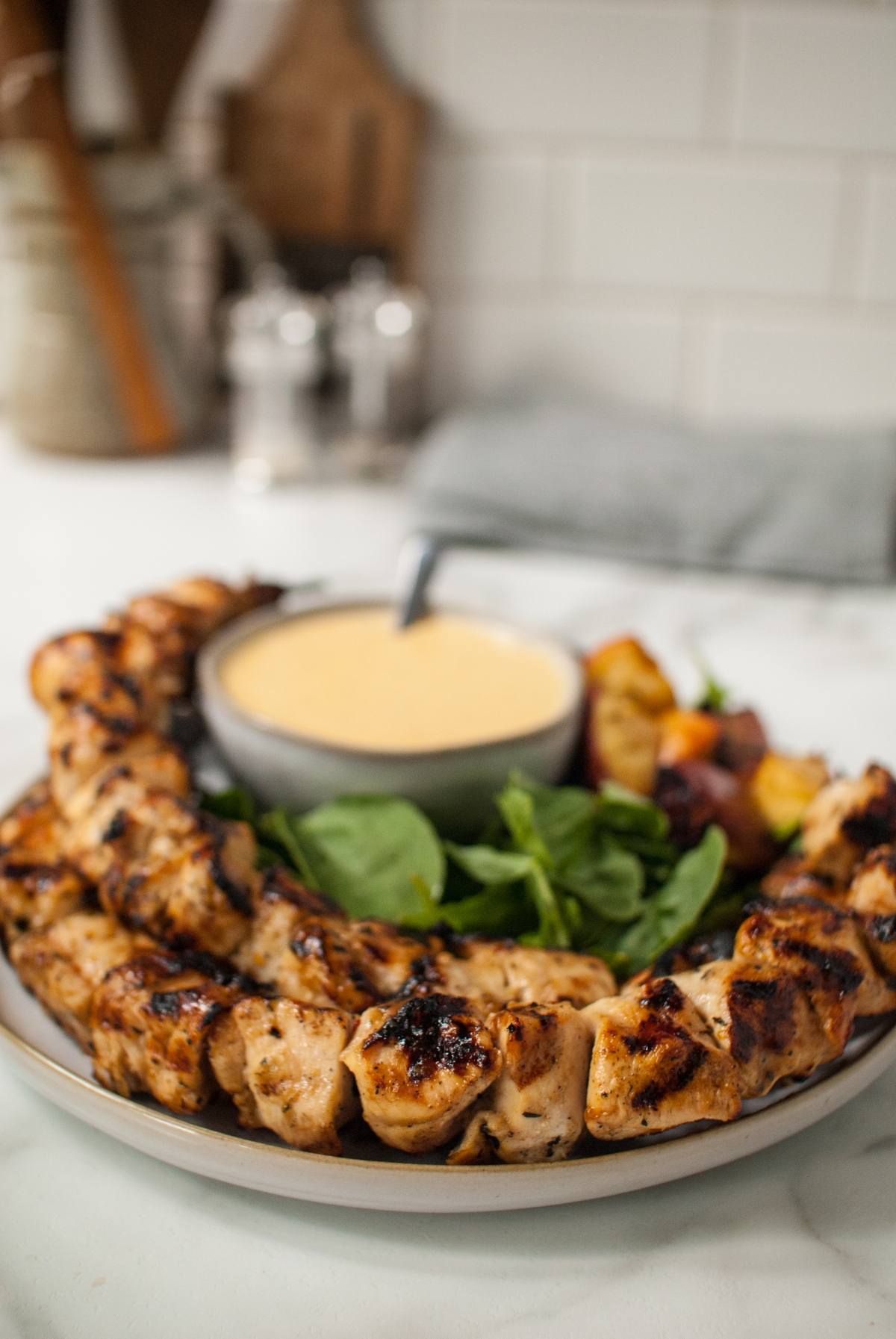 Cider grilled chicken skewers arranged on a plate with fresh greens and a bowl of Gouda cheese sauce for dipping.