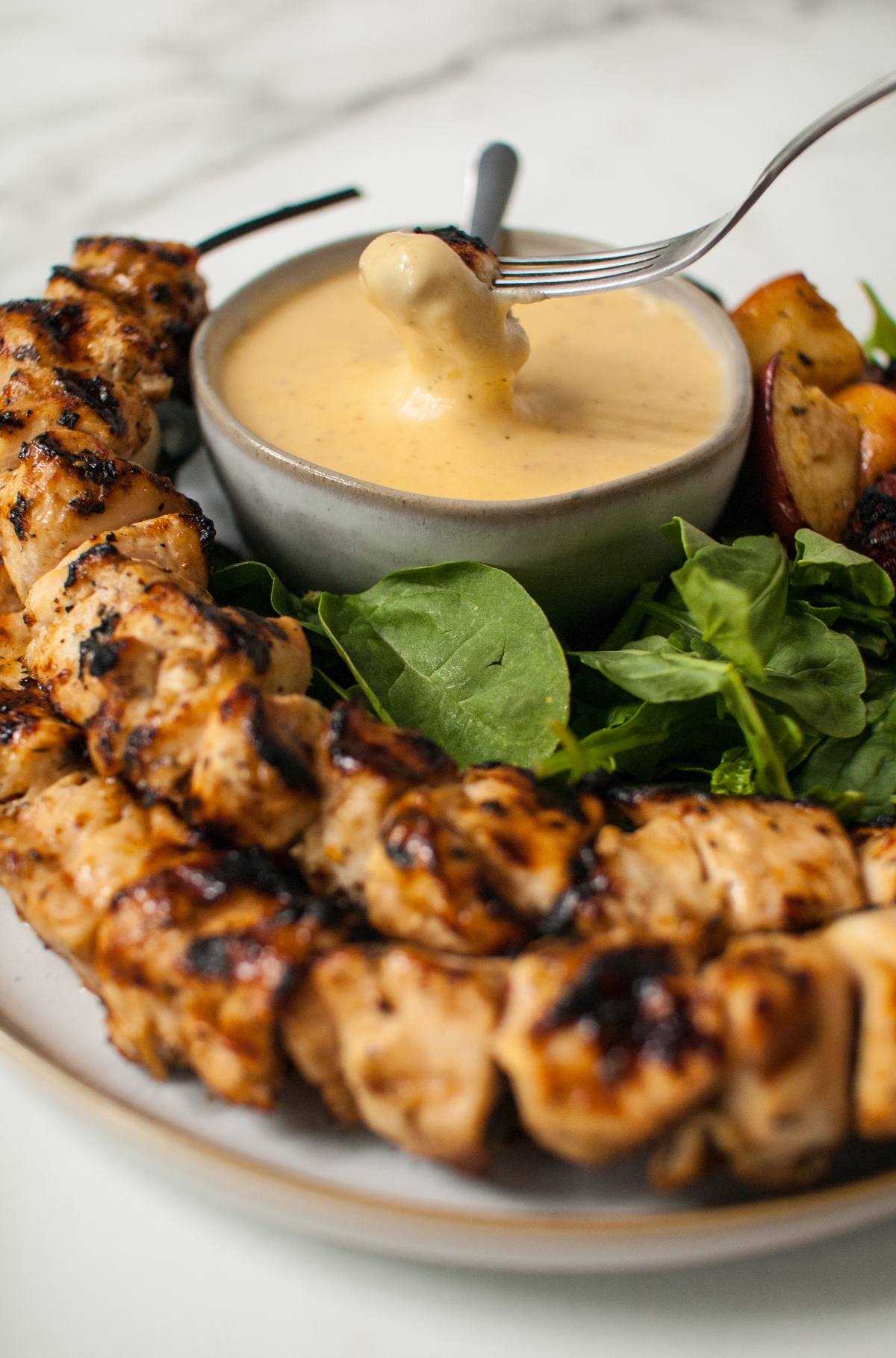 Grilled cider chicken skewers with greens and a fork dipping chicken into a bowl of rich Gouda cheese sauce.