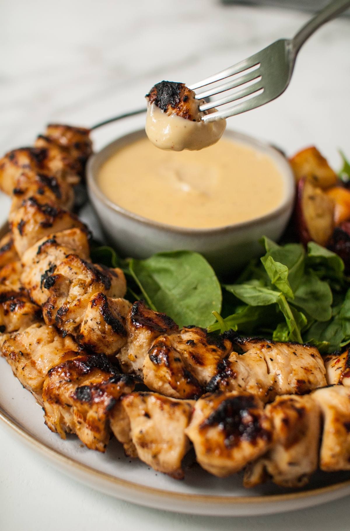 Apple Cider Chicken Skewers with Gouda Sauce; greens on the side and a fork dips tender chicken into a creamy Gouda Cheese Sauce.