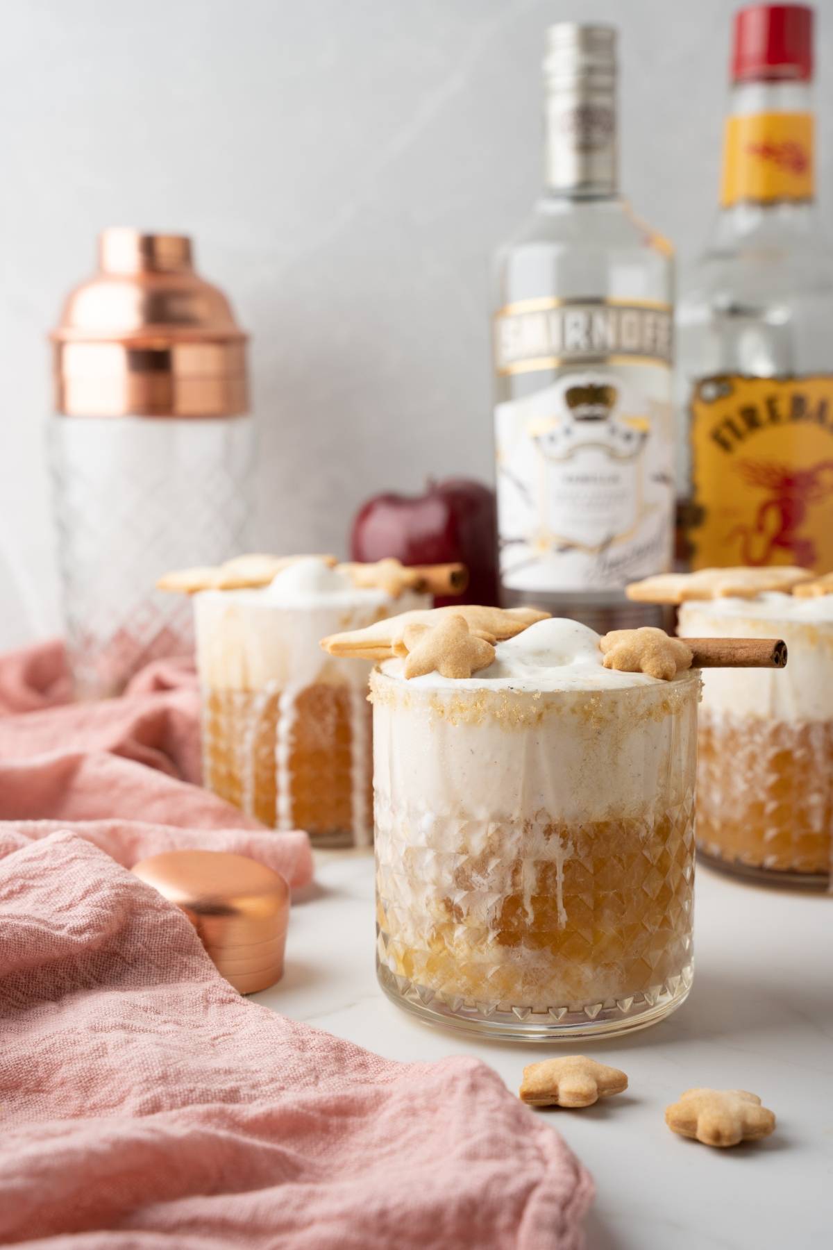 Two cocktails with whipped topping, cinnamon sticks, and cookies are served alongside bottles and a shaker—perfect for trying an autumn cocktail recipe like an Apple Pie Cocktail.