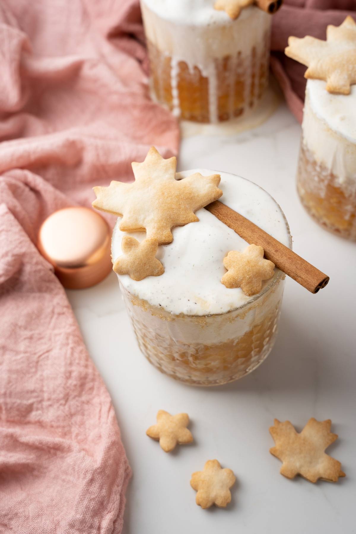A frothy autumn cocktail recipe served in a glass, topped with a maple leaf cookie and cinnamon stick, surrounded by small cookies—an inviting apple cinnamon cocktail perfect for cozy evenings.