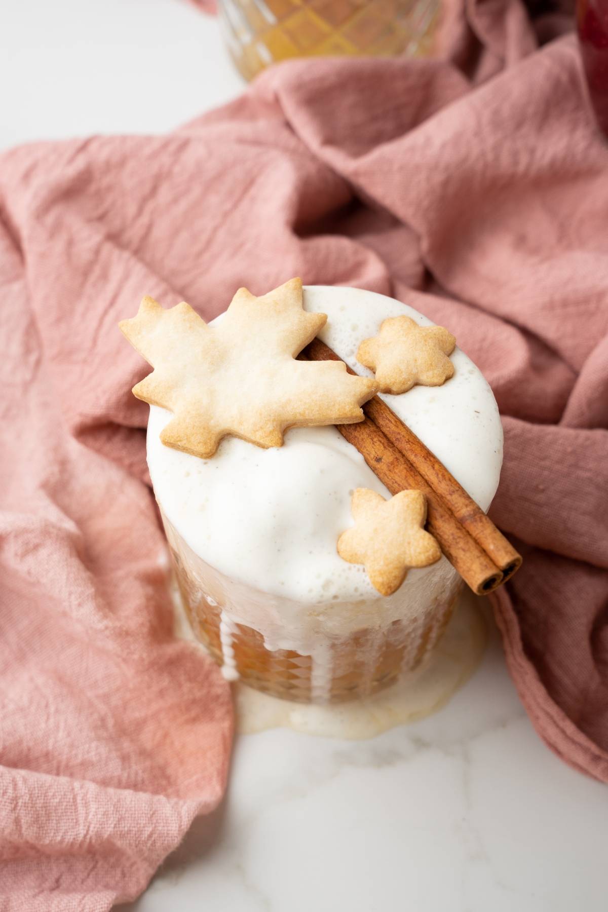A foamy Apple Pie Cocktail topped with a cinnamon stick and leaf-shaped cookies rests on a pink cloth—a delightful autumn cocktail recipe perfect for cozy gatherings.