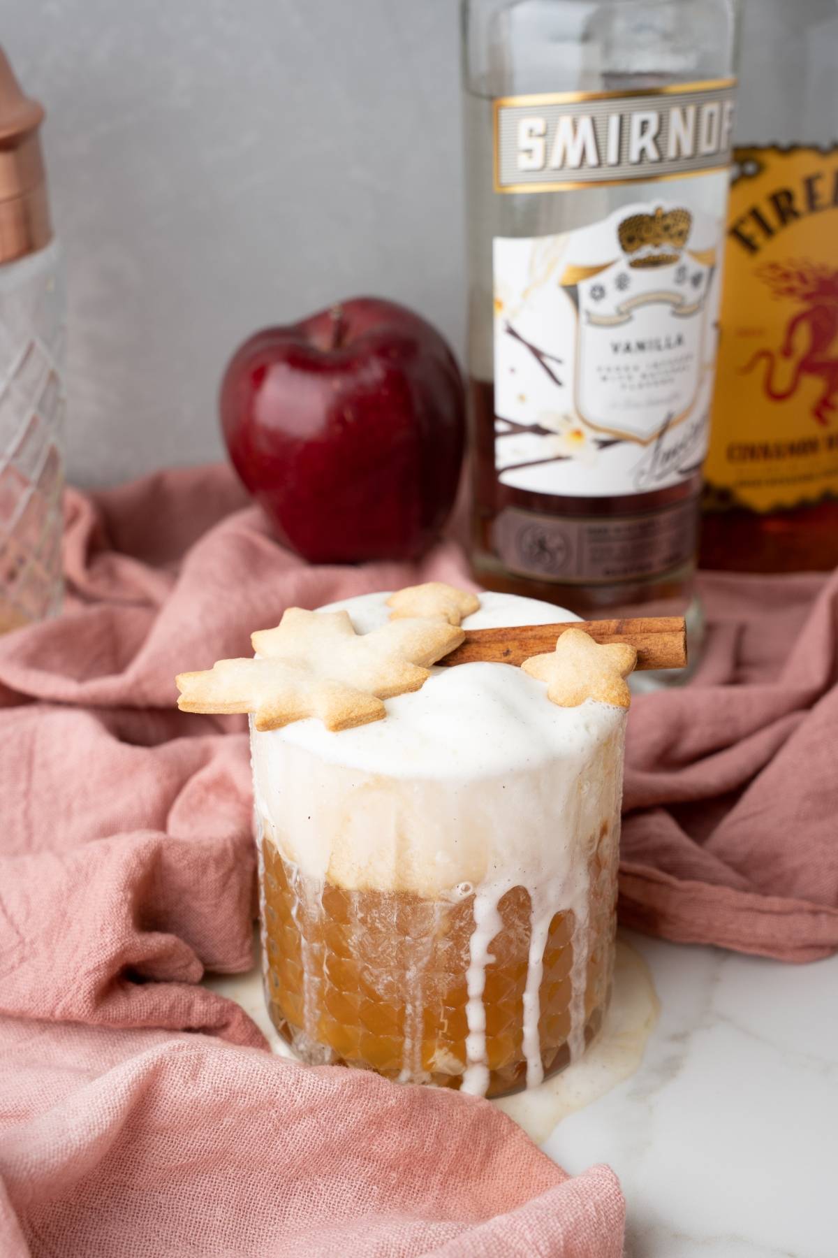 An Apple Pie Cocktail topped with foam, star-shaped cookies, and cinnamon sticks, with bottles and an apple in the background—a perfect autumn cocktail recipe.