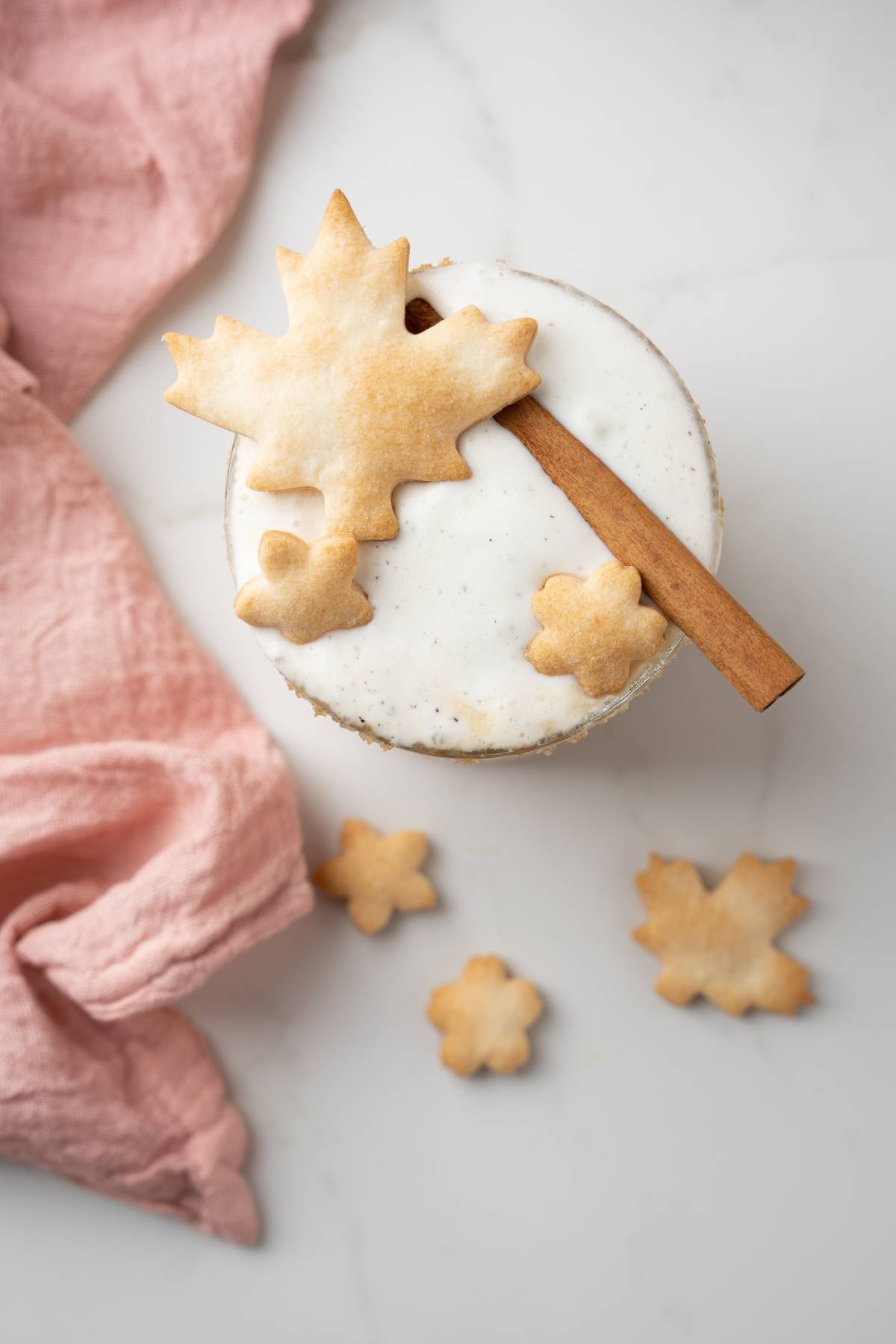 Round cake with white icing, topped with maple leaf cookies and a cinnamon stick—like an Apple Pie Cocktail or cozy autumn cocktail recipe—beside a pink cloth.