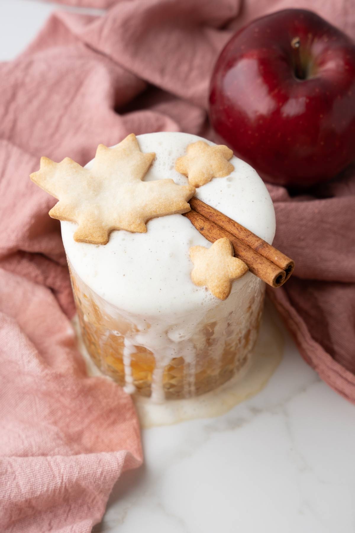 A dessert topped with white icing, leaf-shaped cookies, cinnamon sticks, and a red apple nearby pairs perfectly with an Apple Pie Cocktail.