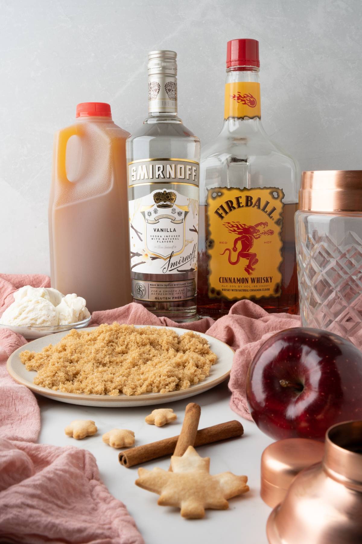 A display of vanilla vodka, Fireball whisky, apple cider, brown sugar, whipped cream, an apple, and cinnamon sticks for an irresistible Apple Pie Cocktail—an autumn cocktail recipe made with apple cider.