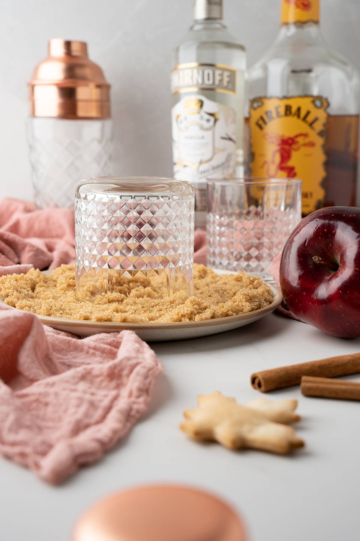 A glass rimmed with sugar sits on a plate, surrounded by bottles, an apple, and cinnamon sticks—perfect for crafting an apple cinnamon cocktail.
