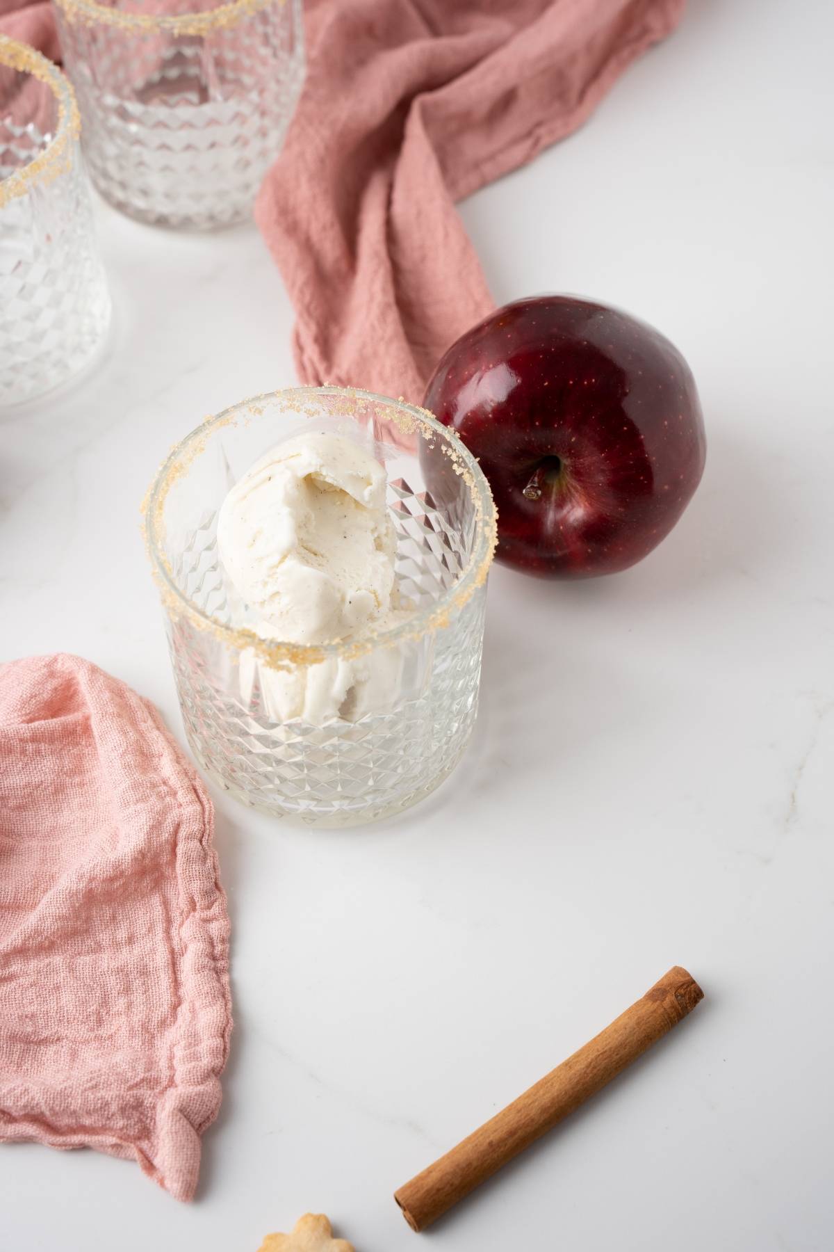 A glass with vanilla ice cream, a red apple, a cinnamon stick, and pink cloth on a white surface—perfect inspiration for an autumn cocktail recipe or a cocktail made with apple cider.