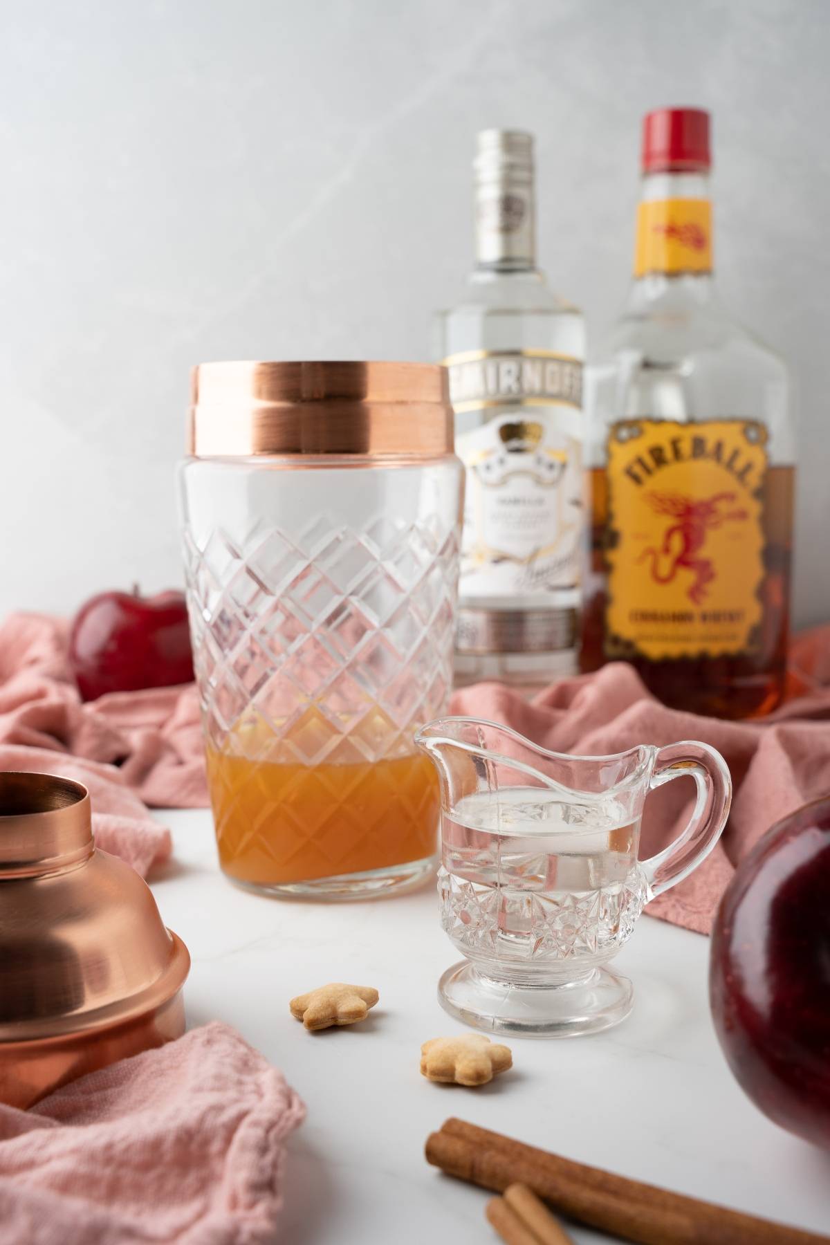 Cocktail shaker, glass of liquid, cinnamon sticks, and bottles of alcohol on a white surface with red cloth—perfect setup for crafting an apple cinnamon cocktail or your favorite autumn cocktail recipe.