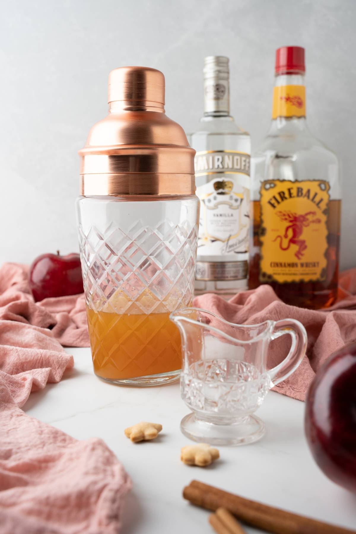 A cocktail shaker, small glass pitcher, liquor bottles, apples, and cinnamon sticks on a white surface hint at an Apple Pie Cocktail—a cozy cocktail made with apple cider and perfect for mixing with Fireball Whisky.