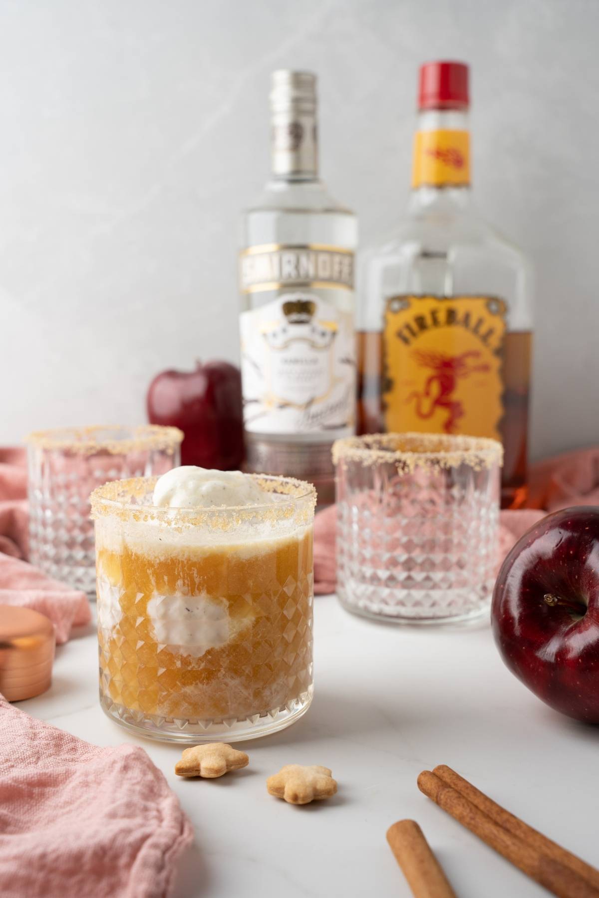 Glass of apple pie cocktail with ice cream, surrounded by apples, cinnamon sticks, and bottles of vodka and Fireball Whisky—a delicious autumn cocktail recipe made with apple cider.